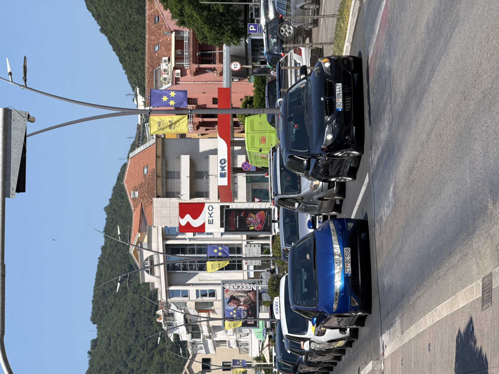 This is a medium-shot, eye-level, outdoor, daytime photograph of a traffic jam on a city street. The street is paved and has lane markings. The buildings in the background are multi-story and appear to be commercial, with red-tiled roofs and a large EKO gas station sign. There are several cars, including a blue Honda Civic in the foreground with license plate CT BC 508, and a black BMW in front of it with license plate BD BN447. Other vehicles visible include a white taxi, a green van, and several other cars. The sky is clear and blue, and there are green hills in the background. This scene is typical of a busy urban environment during peak hours, likely the city of Budva, Montenegro, known for its tourist traffic and congestion during the summer months.