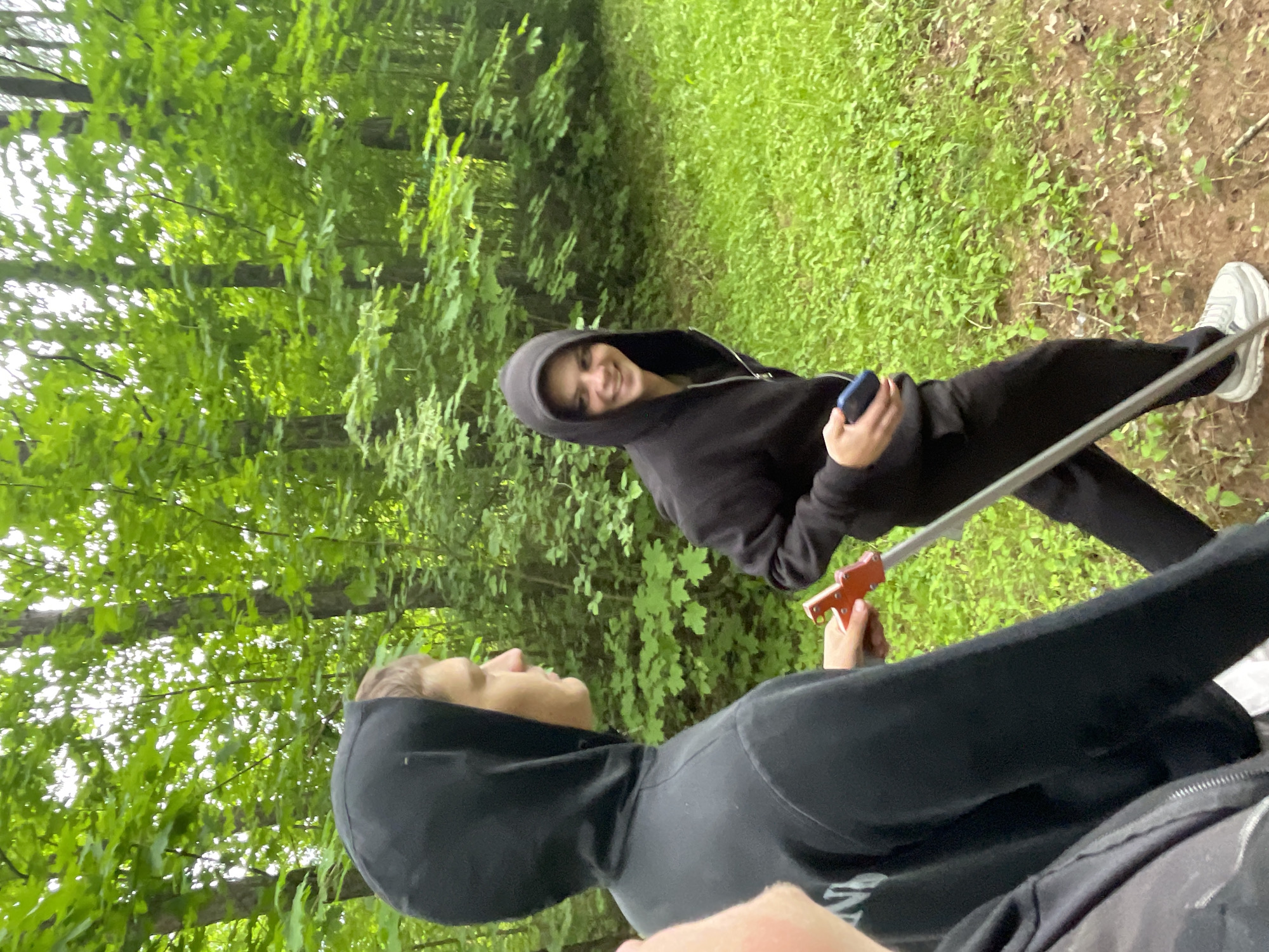 The image, rotated on its side, depicts two individuals in a vibrant, green outdoor setting, likely a forest or wooded area in Красные Баки, Russia. The background is filled with lush green foliage, including tree leaves and dense ground cover, with glimpses of sunlight filtering through the canopy.
One person, positioned further back on the left, is wearing a black zipped hoodie, black pants, and white athletic shoes. They are holding a blue smartphone in their left hand and are looking directly at the camera with a clear smile, suggesting a relaxed and positive mood.
In the foreground, closer to the right, a second individual is partially visible, seen mostly from the side and back. This person is also wearing a black hoodie and appears to be looking upwards or towards the first individual. Their right hand is holding a small, red object, which resembles a toy or a simple tool. A light-colored pole or stick extends diagonally across the lower center of the frame, its purpose unclear but possibly part of outdoor equipment.
The bright lighting indicates that the photograph was taken during the daytime. There is no visible text in the image.