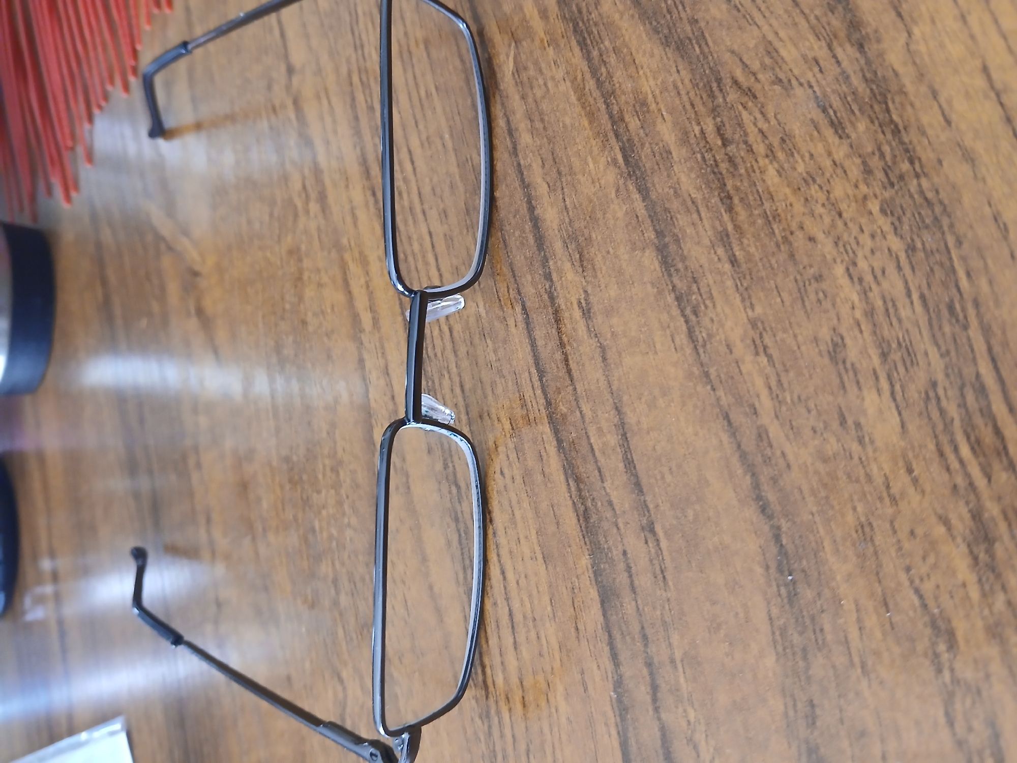 A pair of black-rimmed eyeglasses with clear lenses are resting on a brown wooden surface. The glasses are positioned horizontally across the frame, with the temples extending outwards. The wooden surface has a visible grain pattern. The background is blurred, but appears to show a dark circular object and some red, thin objects. The scene is indoors, likely on a table. The location is not specified to be in Moscow, Russia.