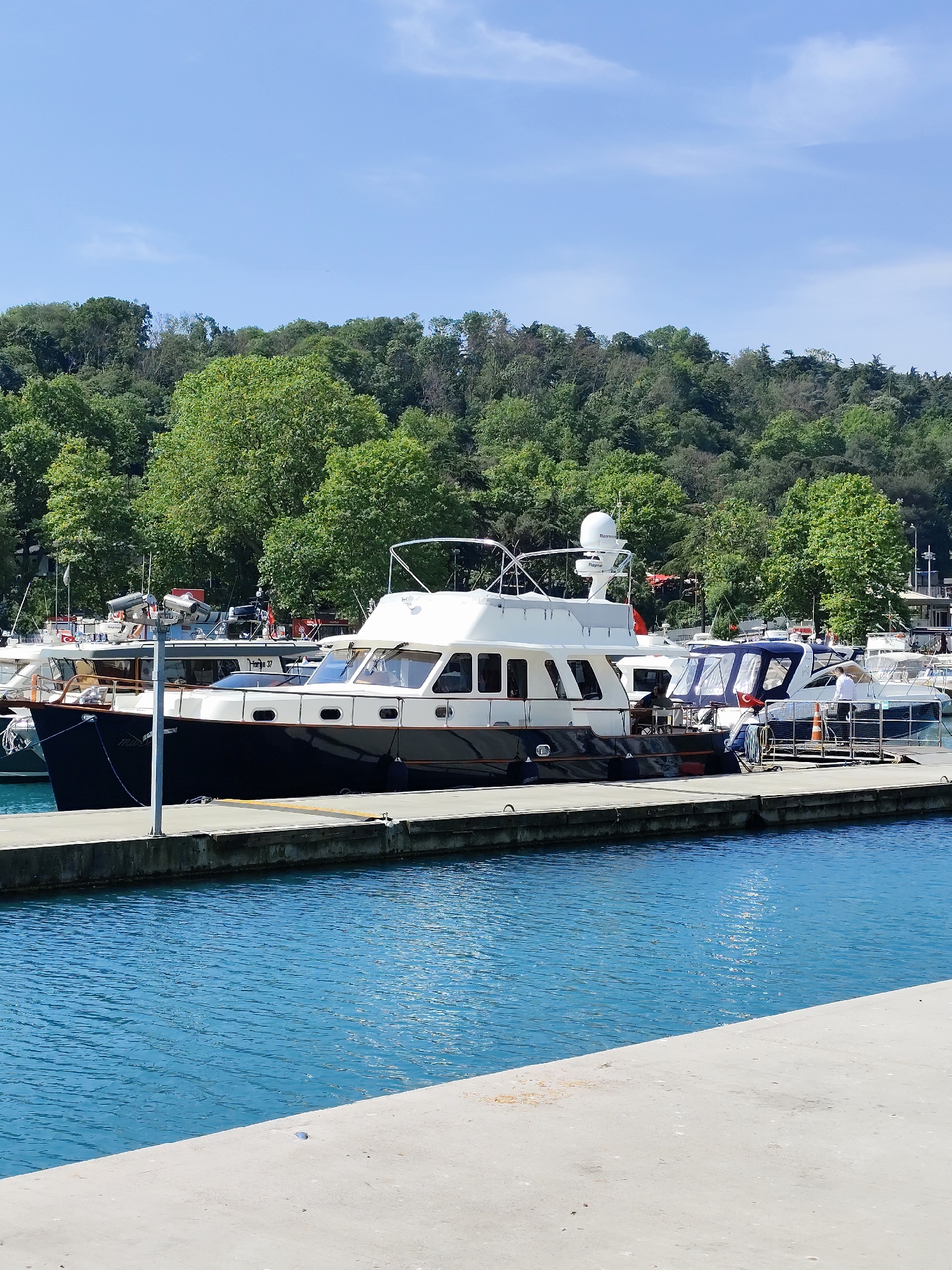The image shows a marina in Sarıyer, Turkey on a clear day. A large, two-story motor yacht with a dark blue hull and white cabin is docked alongside a concrete pier. A smaller motorboat is docked next to it. The surrounding environment includes a forested hillside in the background and clear turquoise water.