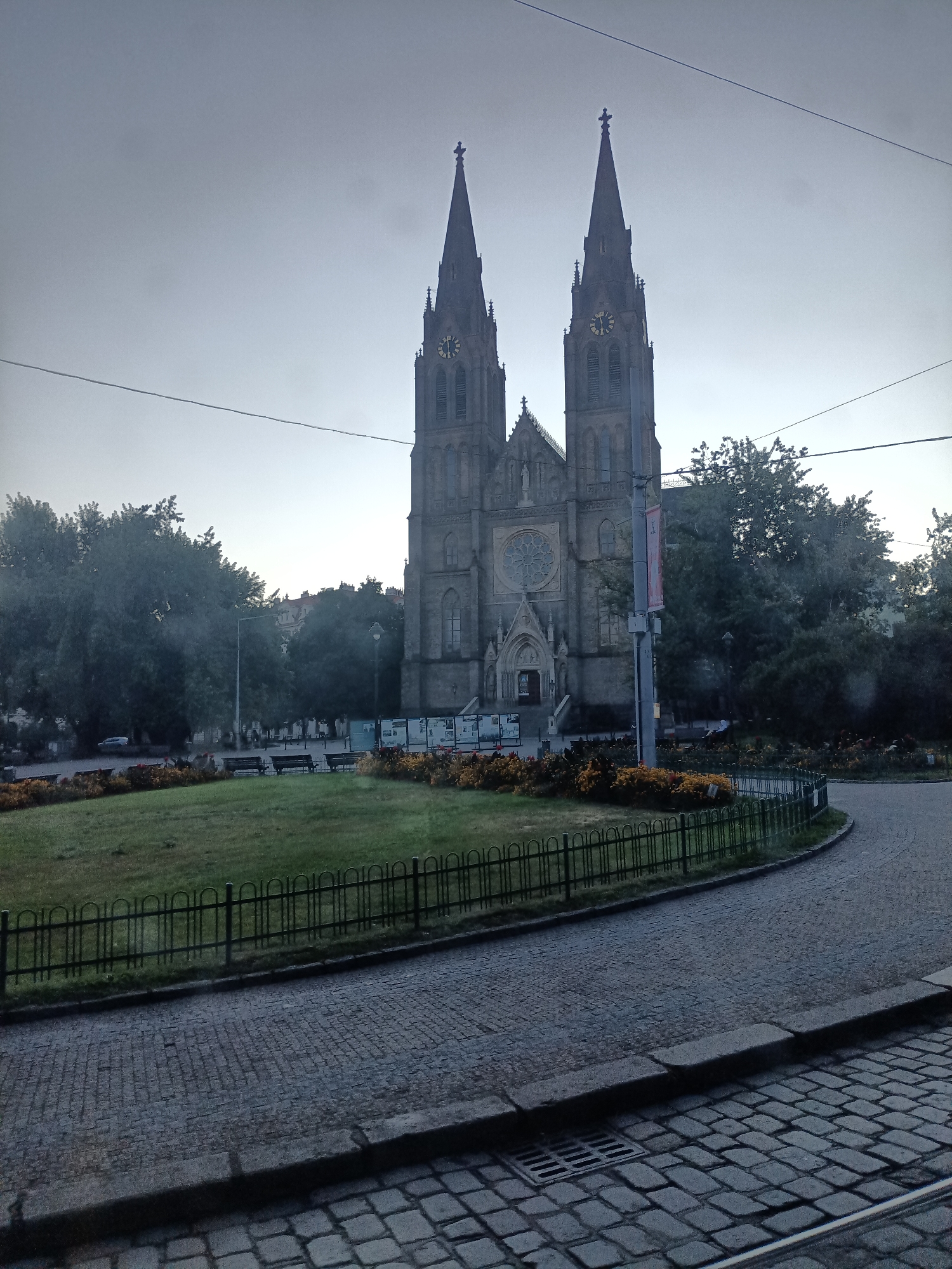 This is a full, eye-level shot of the Basilica of Saint Peter and Saint Paul in Prague, Czechia. The basilica is a large, Gothic Revival structure with two prominent spires and a rose window above the entrance. The facade is made of grey stone, and clocks are visible on each spire. The basilica is framed by trees and a public park in the foreground. A cobblestone street curves around the park, and a low black fence encloses the green space. The sky is overcast and grey, suggesting it is either early morning or late evening, or simply a cloudy day. There are no people visible in the image, but there are several benches in the park. Electrical wires are visible crisscrossing the sky in the upper portion of the image.