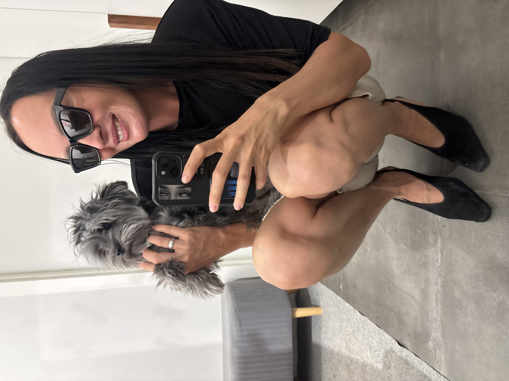 A woman is taking a selfie with a small, gray, fluffy dog. She is crouching and wearing a black t-shirt, beige shorts, sunglasses, and black flats. She is holding a black smartphone with a picture of eyes on the case. The woman has long dark hair and is smiling. She is wearing a ring on her finger. The background appears to be a room with white walls and a gray floor. There is a gray stool with a wooden leg visible in the lower portion of the image. The location of the photograph is in Chaikovsky, Russia.