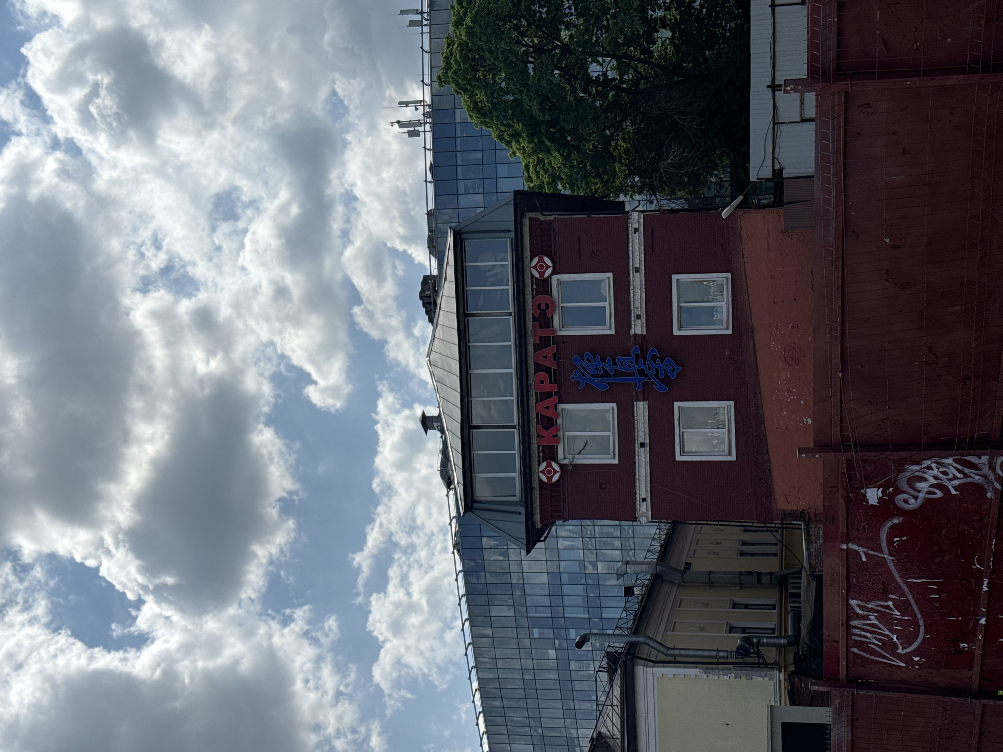 A medium shot of a four-story brick building with a sign that reads "KARATE" in Russian, located in Moscow, Russia. The building has white-framed windows and a gray roof with a slanted section. To the left of the building is a modern glass building that reflects the sky. The sky is visible in the background with scattered white clouds. To the right of the brick building is a red wall with graffiti. A tree is partially visible above the red wall and glass building. The image is taken from a low angle.