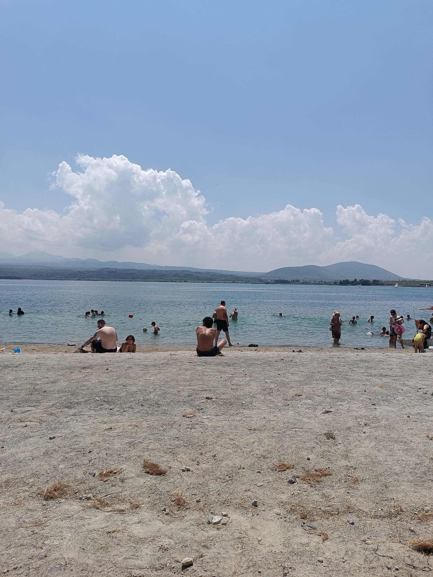A wide shot captures a sunlit beach scene on the shore of Lake Sevan, Armenia. The foreground is dominated by sandy ground with scattered dry grass and small stones. Beyond the beach, a large body of calm, blue water is populated with numerous people swimming and wading. In the middle ground, several individuals are seated on the sand closer to the water, some of whom appear to be observing those in the lake. In the background, rolling green hills and mountains form a distant shoreline, under a bright blue sky with scattered fluffy white clouds. The overall atmosphere is one of a leisurely summer day at a popular lakeside recreational area.