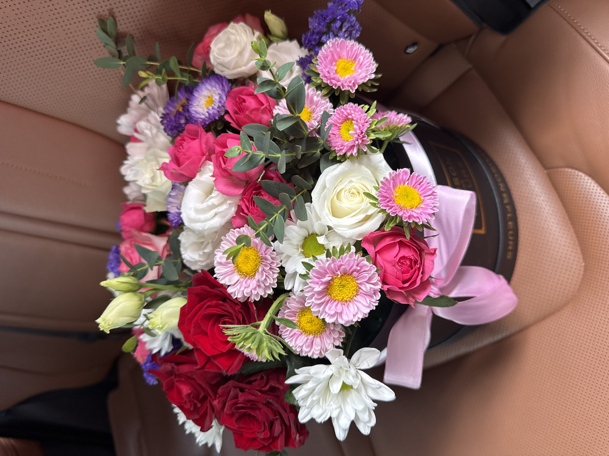 The image shows a bouquet of flowers in a dark circular box, tied with a pink ribbon, placed on a tan leather car seat. The bouquet contains a variety of flowers, including red roses, white roses, pink roses, white chrysanthemums, pink asters with yellow centers, purple asters, and light blue flowers. Green eucalyptus leaves are also interspersed throughout the bouquet. The car interior appears to be luxurious, with perforated leather upholstery visible. The lighting suggests it is daytime. The specific location in Almaty, Kazakhstan cannot be determined from the image alone.