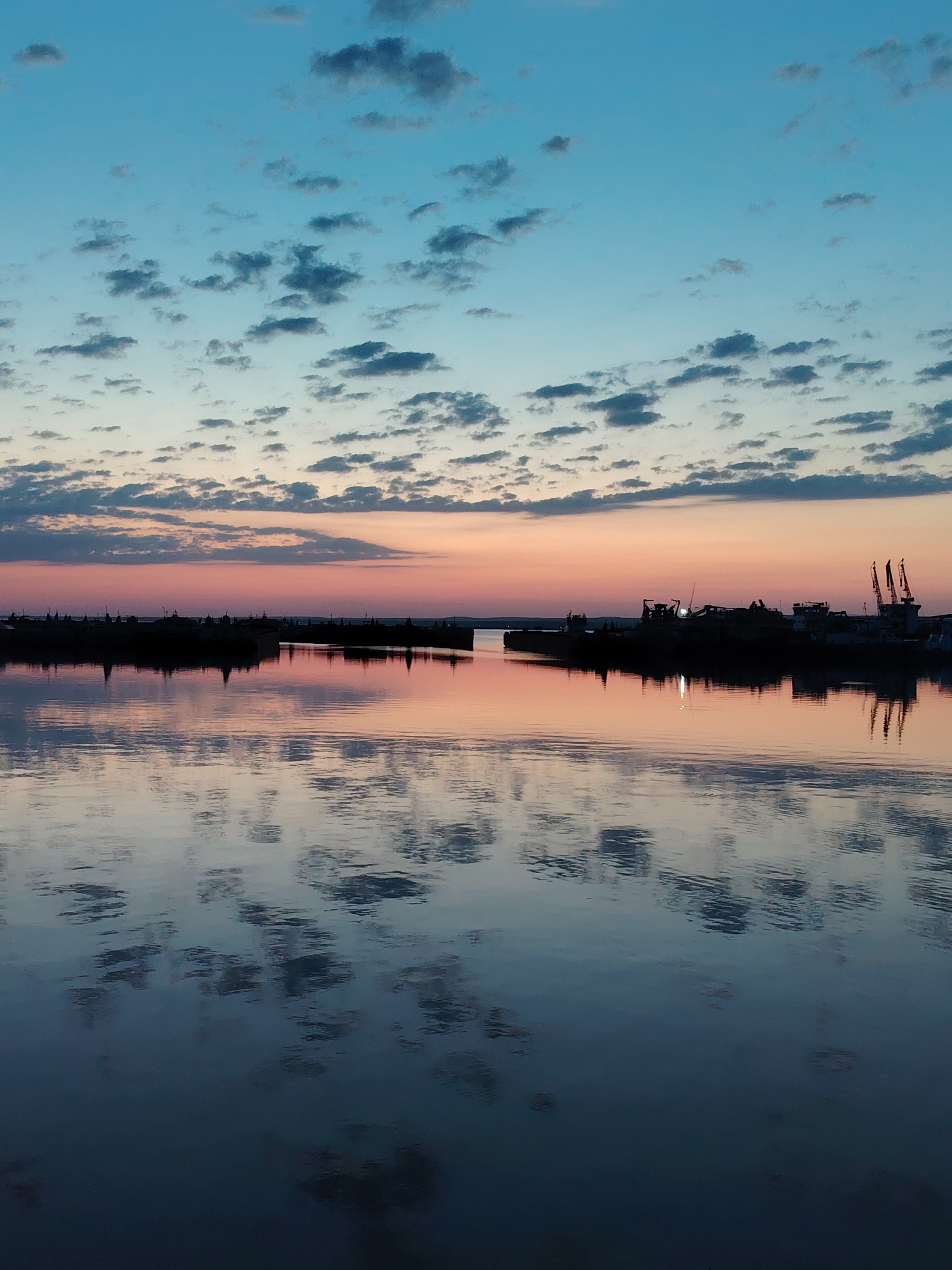 The image depicts a tranquil scene at sunset in Chaikovsky, Russia. The sky transitions from blue to pink and orange hues, dotted with small, scattered clouds. The water reflects the sky and clouds, creating a mirror-like effect. Silhouetted against the horizon are structures, potentially boats and cranes, within what appears to be a port or industrial area along the water's edge. The overall mood is calm and serene, capturing the stillness of the evening. There is no evidence of any people.