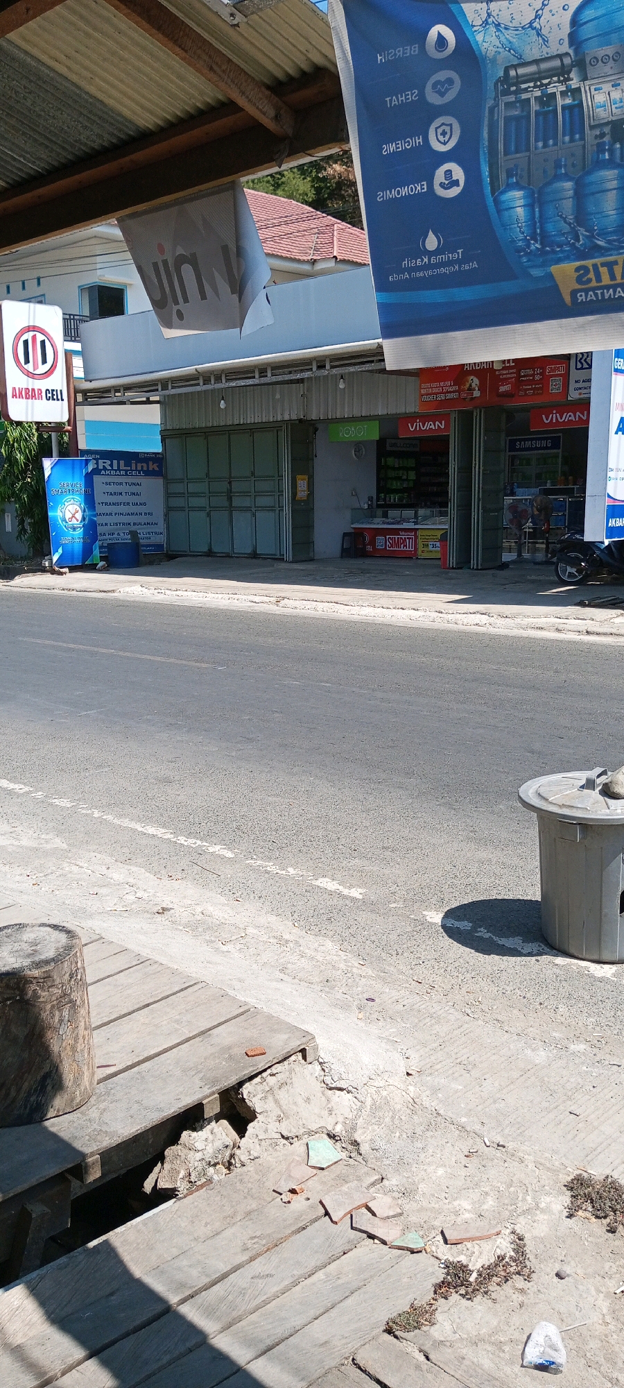 The image shows a street scene in Sengkang, Indonesia, featuring a row of shops. In the foreground, a wooden walkway is visible, partially broken and showing debris underneath. A large tree stump stands on the left side of the walkway. To the right, a gray cylindrical trash can is situated near the roadside.
The shops are identified by various signs. "AKBAR CELL" is advertised on a vertical sign near the left edge of the frame. A large banner displaying water bottles and text in Indonesian, possibly related to water purification or sales, hangs overhead and partially obscures the view of the shops. Another banner with the "vIn" logo is also hanging and torn.
The shops themselves appear to be small retail establishments. One shop has a large glass-paneled folding door. Another visible shop displays "VIVAN" and "SAMSUNG" branding. Inside the Vivan shop, shelves are stocked with various products, and a counter is present. A "ROBOT" sign is also visible above one of the shop entrances. A motorbike is parked in front of one of the shops.
The street in front of the shops has a white dashed line marking the lane. The overall environment suggests a commercial area in Sengkang, with buildings lining the street. The time of day appears to be sunny, with clear shadows cast by the objects.