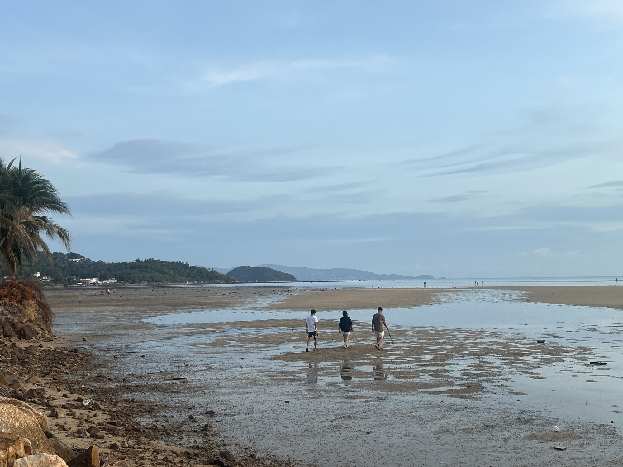 The image shows a coastal scene in Baan Sri Thanu, Thailand. A wide, shallow tidal flat dominates the foreground. The receding tide has left pools of water on the sand. Three people are walking away from the viewer on the flats. From left to right, the first person is wearing a white shirt and shorts, the second a dark top and light shorts, and the third a dark shirt and light shorts while holding a long stick. In the background, a coastline features low hills and sparse development of buildings visible along the tree line. Mountains can be seen further inland, shrouded in atmospheric haze. The sky is overcast with blue and grey clouds. Rocks and a palm tree are in the lower left corner.