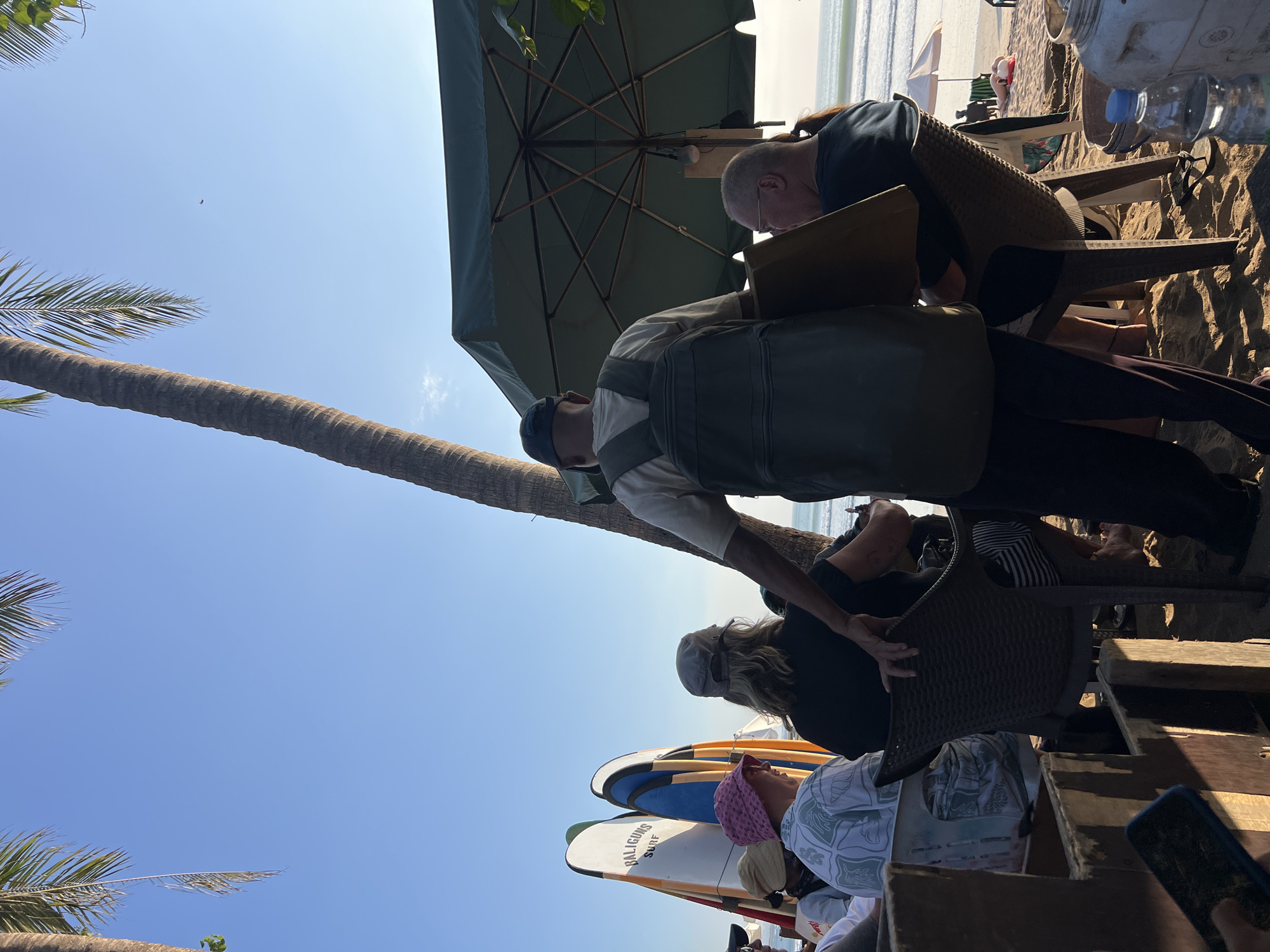 In Kuta, Indonesia, at a beachside establishment, several individuals are seated at tables on the sand. A tall palm tree looms over the scene, casting shadows. In the background, surfboards are propped up. A server, wearing a backpack, interacts with a seated patron. The ocean is visible in the distance. The atmosphere suggests a casual dining or relaxation area on the beach.