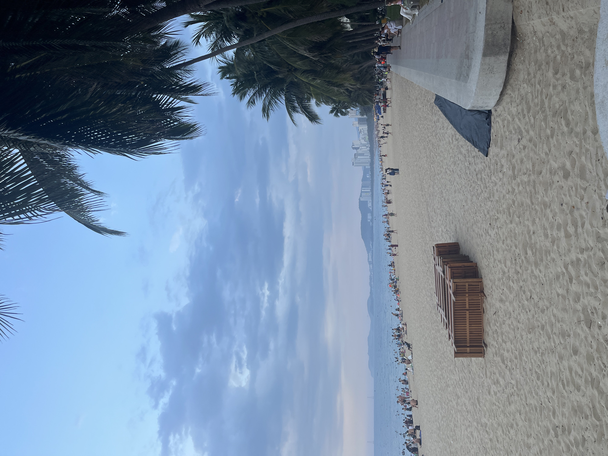 The image depicts a beach scene in Nha Trang, Vietnam, under an overcast sky. The foreground is dominated by sand, with a stack of wooden lounge chairs placed towards the left. A dark tarp is visible on the sand near a concrete walkway that runs along the right side of the beach.
Further back, the beach is populated with numerous people, many of whom are seated or standing on the sand, and some are in the shallow water. The ocean stretches to the horizon, with a faint line of hills and a cityscape visible in the distance. Palm trees line the right side of the beach, framing the view of the ocean and city. The overall atmosphere suggests a busy, public beach during daylight hours, possibly late afternoon or early evening given the muted light.