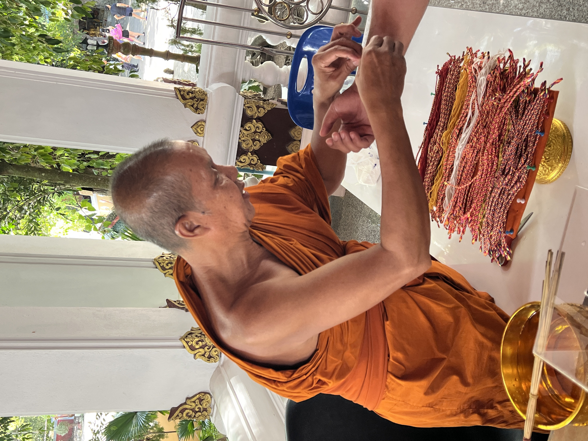 In Baan Hua Thanon, Thailand, a Buddhist monk, wearing an orange robe, is shown tying a red and white braided string bracelet around a person's wrist. The monk's head is shaved and he is looking down at his task. In front of him on a white table is a golden bowl with water and sticks, and a stand displaying a large pile of similar braided bracelets in red, yellow, and white colors. Behind the monk, pillars with ornate gold carvings and a railing are visible, with trees and other people in the background, suggesting an outdoor setting at a temple or similar religious site.
