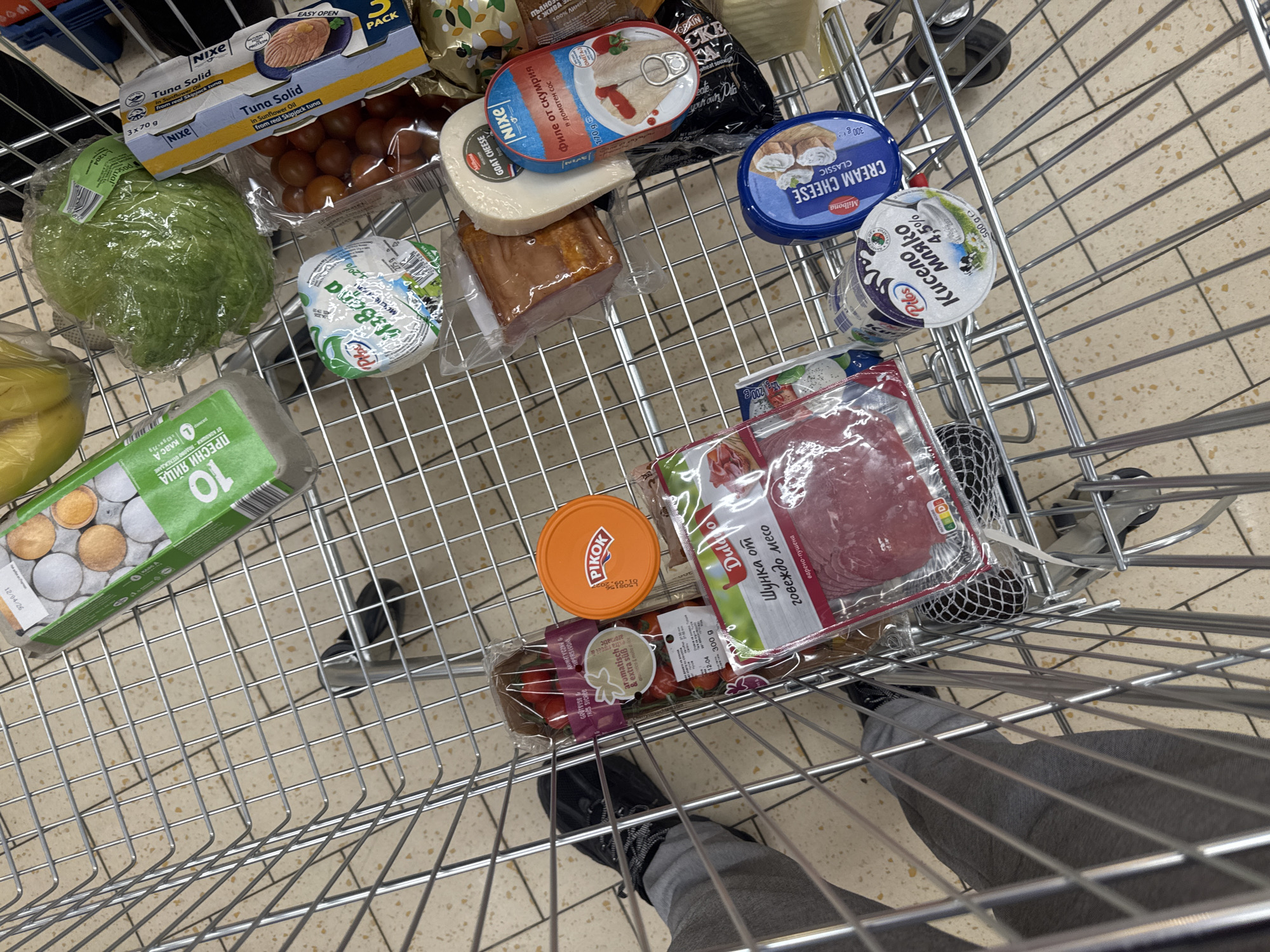 A shopping cart in Sofia, Bulgaria, contains grocery items: a Nixe Tuna Solid 3-pack, tomatoes, a head of lettuce, a package of eggs, Milbona Cream Cheese, goat cheese, canned fish, a container of yogurt, sausage slices, and Pikok liver pate. The shopper's legs and feet in black shoes are visible near the bottom of the frame.