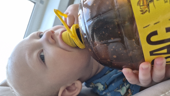 An infant is depicted in a close-up, indoor setting within the city of Chaikovsky, Russia. The subject, observed from the chest up, presents with pale skin, sparse light-colored hair, and blue eyes directed slightly upward to the right. Its mouth is actively engaged with the bright yellow plastic sport-cap of a large, clear, ribbed plastic bottle. The bottle contains a dark brown liquid with numerous light-colored suspended particles. A yellow label affixed to the bottle displays partial black Cyrillic text, including "БАС," and an illustration of a wooden barrel with a tap. The infant's left hand is wrapped around and gripping the body of the bottle. The infant is dressed in a blue garment patterned with light-colored cartoon figures. The background is brightly illuminated and out of focus, consistent with a window as the light source.