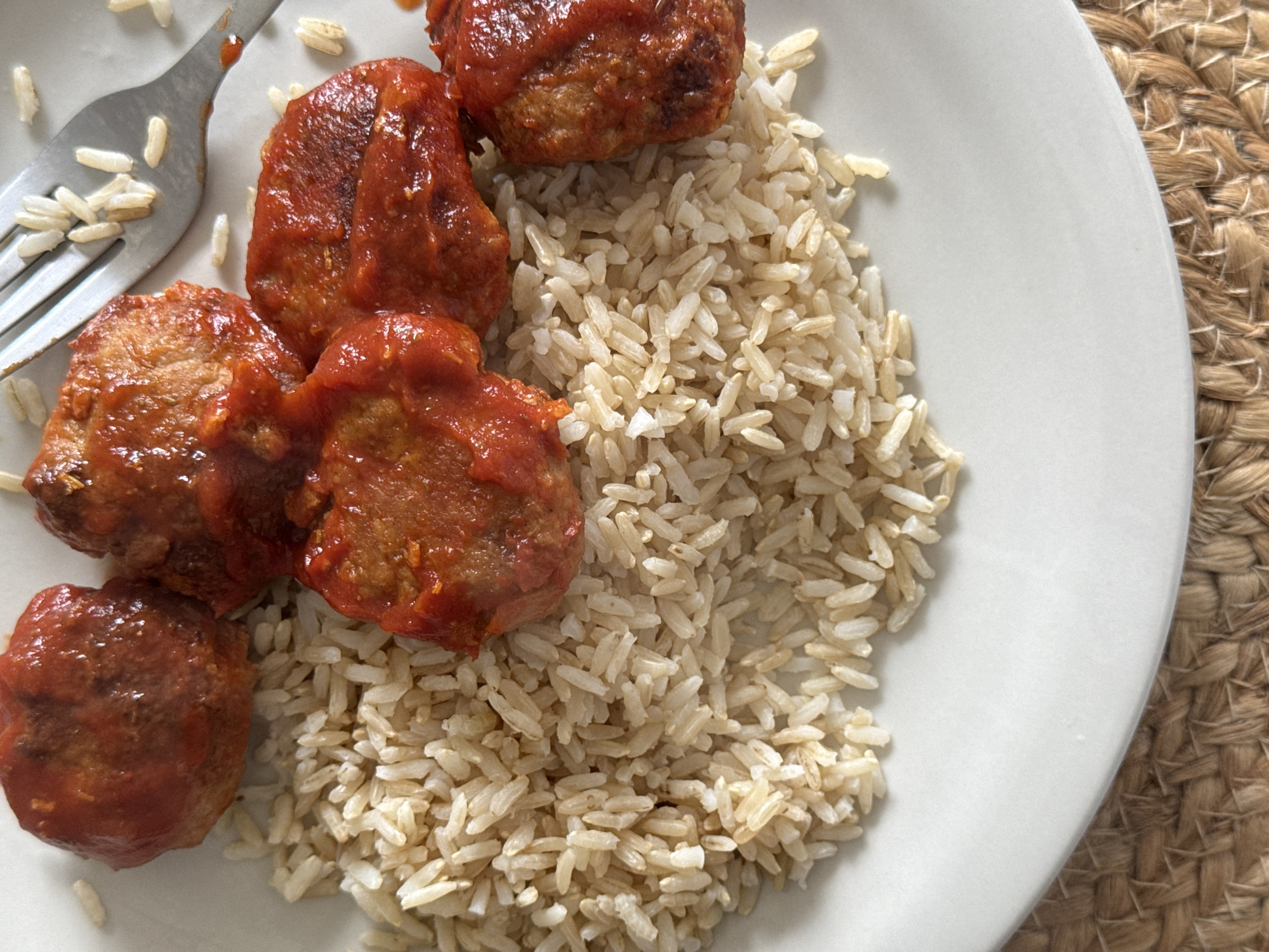 The image shows a plate of food consisting of brown rice and meatballs covered in tomato sauce. There are five meatballs visible, arranged near the rice. A fork rests on the plate beside the food, with a few grains of rice stuck to it. The plate is white and sits on a woven, neutral-toned surface. The location is not discernible from the image.