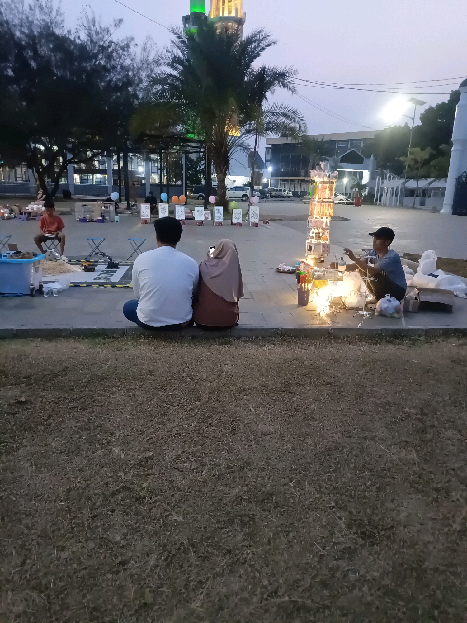 The image depicts an outdoor scene in the city of Lubuklinggau, Indonesia, likely at dusk given the lighting conditions. In the foreground, a couple is seated on the ground facing away from the camera, with the man in a white long-sleeved shirt and the woman in a tan hijab and dress. Adjacent to them, a street vendor is seated on the ground, surrounded by merchandise, including a tower-like display of items illuminated by string lights. The vendor appears to be interacting with the merchandise.
In the middle ground, a young boy in an orange shirt is sitting on a stool. Several small tables with items are set up, along with some display boards featuring drawings of animals. Balloons in various colors are visible behind these displays. In the background, a large building with a distinct green-topped tower, resembling a mosque or religious building, is partially visible. Palm trees and other foliage are scattered throughout the scene. The ground in the immediate foreground is covered in dry grass, transitioning to a paved area where the people and stalls are located. Beyond the paved area, more buildings, trees, and vehicles can be seen. The overall atmosphere suggests a public space, possibly a park or plaza, where vendors have set up shop in the evening.
