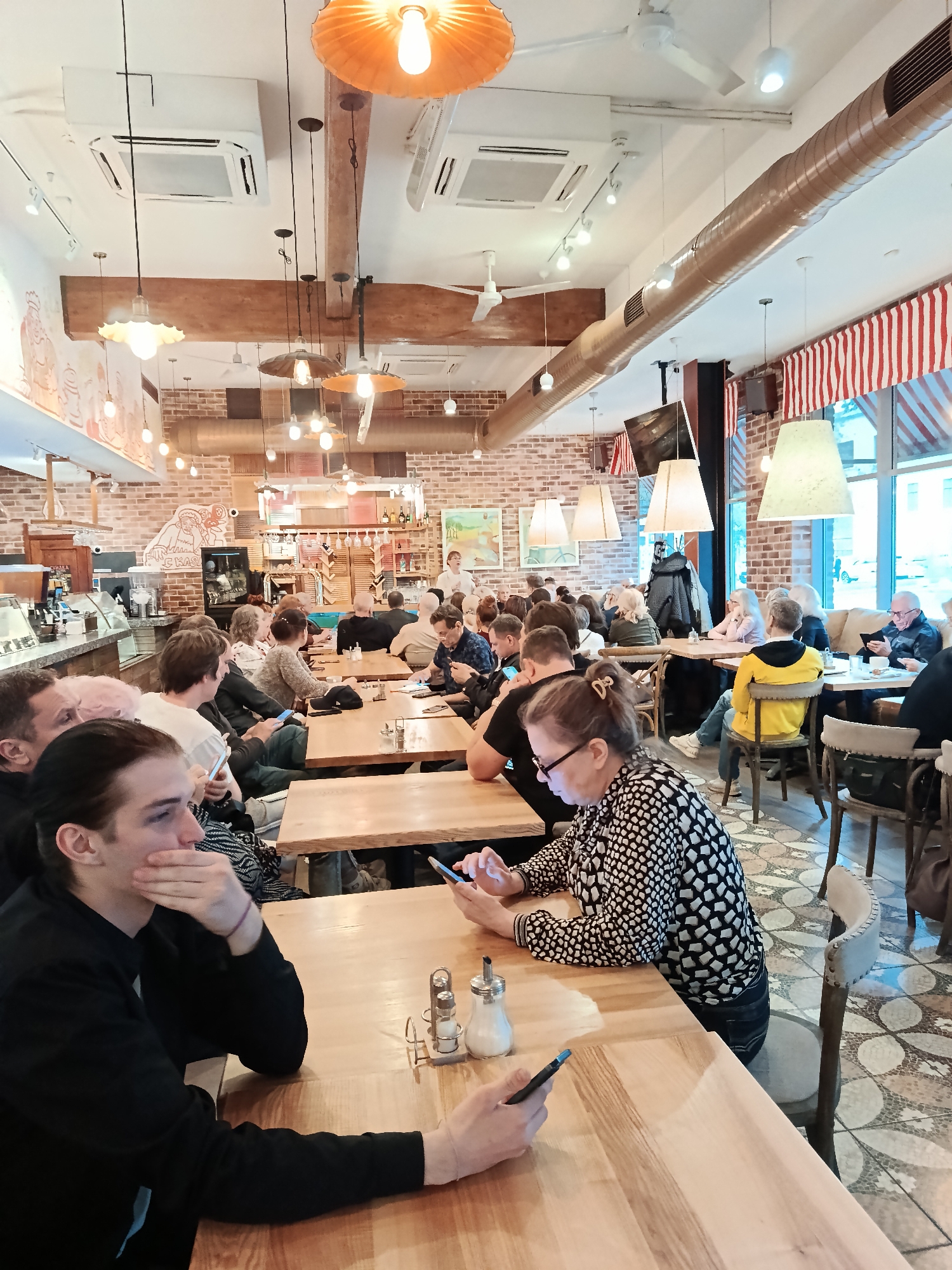 A wide-angle, interior shot depicts a crowded dining room in what appears to be a restaurant. The location is determined to be in Moscow, Russia, based on the red-and-white striped awning and wall decor. A large group of people are seated at wooden tables, most engaged with their phones or looking toward a speaker who stands near the back.
The restaurant features a brick wall backdrop adorned with murals. Overhead, various light fixtures hang, including large orange lamps and smaller industrial-style pendants. Air conditioning units are also visible on the ceiling. The floor is tiled in a mosaic pattern.
The people are of varying ages and dressed in casual attire. Many are looking at their phones.