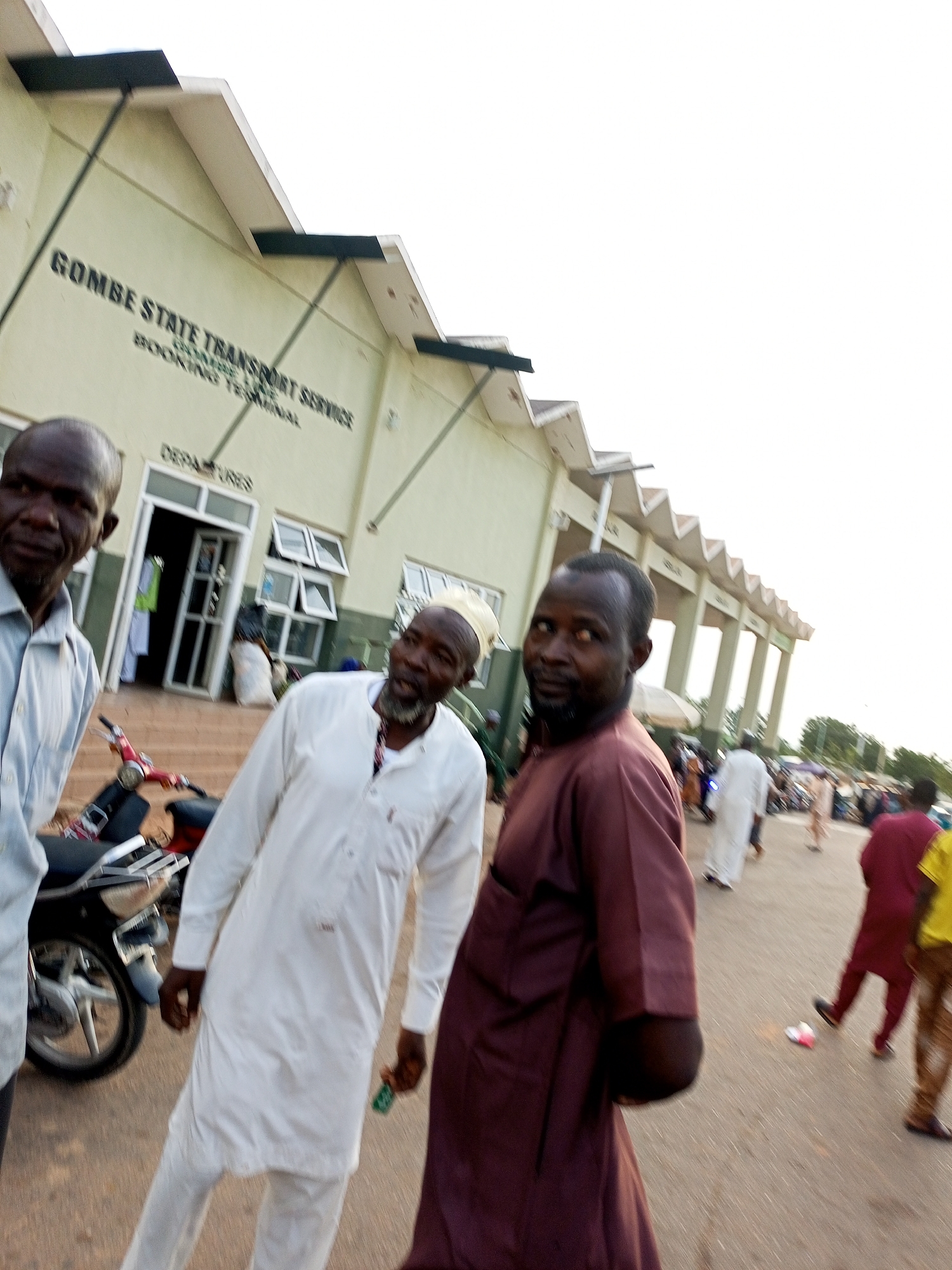 The image depicts the exterior of the Gombe State Transport Service Booking Terminal in Gombe, Nigeria. Three men are prominently featured in the foreground, positioned in front of the terminal building. The man on the left, wearing a light blue collared shirt, is facing the camera with a neutral expression. To his right, a man in a white traditional robe and cap looks towards the camera with an open mouth, appearing to be speaking. The third man in the foreground, on the far right, is seen from the side and back, wearing a maroon garment. He is looking over his shoulder towards the left of the frame.
Behind the men, the transport terminal building is visible, with a light green facade and a textured roofline. Text on the building reads "GOMBE STATE TRANSPORT SERVICE" and "BOOKING TERMINAL". A doorway with glass panels and a "DEPARTURES" sign is present on the building's facade. Several windows are also visible.
In the middle ground, a red and black motorcycle is parked. Further into the background, other individuals can be seen, along with some market stalls or makeshift structures, suggesting a bustling environment. The ground is paved and appears to be a roadway or a large open area. The lighting suggests it is daytime, possibly late afternoon or early evening. The overall scene captures a moment at a transportation hub in Gombe.