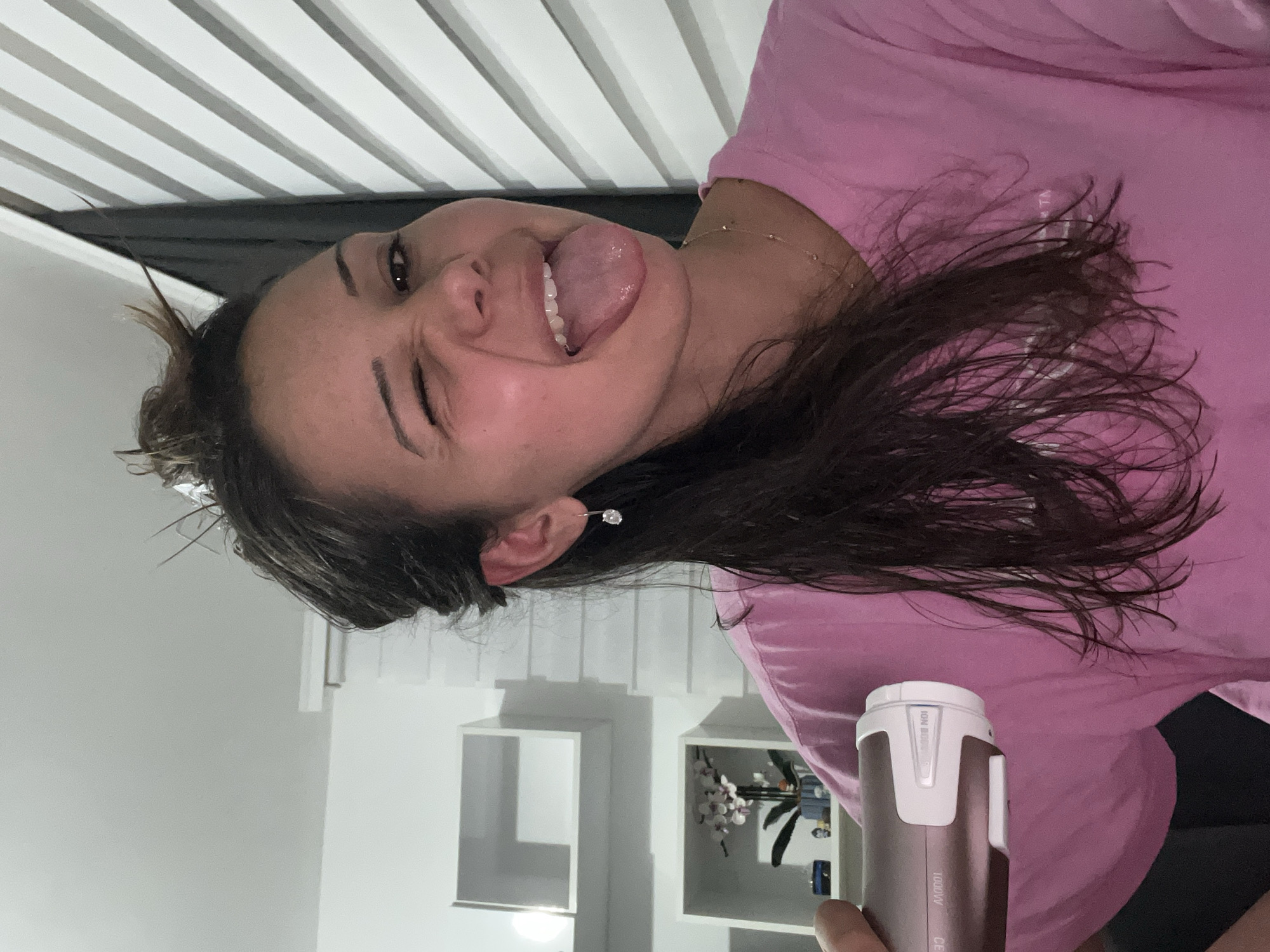 An indoor, head-and-shoulders portrait captures a young adult female in the city of Zlatograd, Bulgaria. She is positioned slightly off-center, facing the camera with a playful expression, winking with her right eye and sticking her tongue out. Her dark, wet hair is partially pulled up in a loose bun on the top of her head, with strands falling over her shoulders and chest. She wears a pink long-sleeved t-shirt, a delicate golden necklace, and a single silver drop earring with a clear stone in her visible left ear. In her left hand, partially visible on the lower left, she holds a rose-gold colored hair dryer with white accents, displaying text including "1000W" and "ION BOOST". The background on the left features a white wall with two rectangular, open white wall shelves; the lower shelf holds a vase with artificial white and pink flowers. On the right, white horizontal blinds are visible. The scene is brightly lit by indoor artificial lighting.