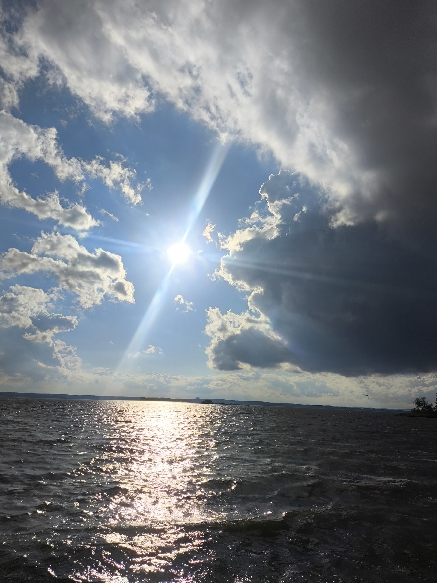 This is a medium-angle, low-light, outdoor, vertical, handheld photograph of a large body of water, possibly a lake or reservoir, under a dramatic sky. The sun is bright and low in the sky, casting a strong glare across the rippling water in the foreground. There are scattered white clouds against a bright blue sky, with a large, dark storm cloud dominating the right half of the sky. The horizon line is visible in the distance, with faint landmasses and a few scattered structures. The overall impression is one of dynamic weather conditions, with a mix of sunlight and impending rain. The location is identified as Zhdanovichi rural council, Belarus.