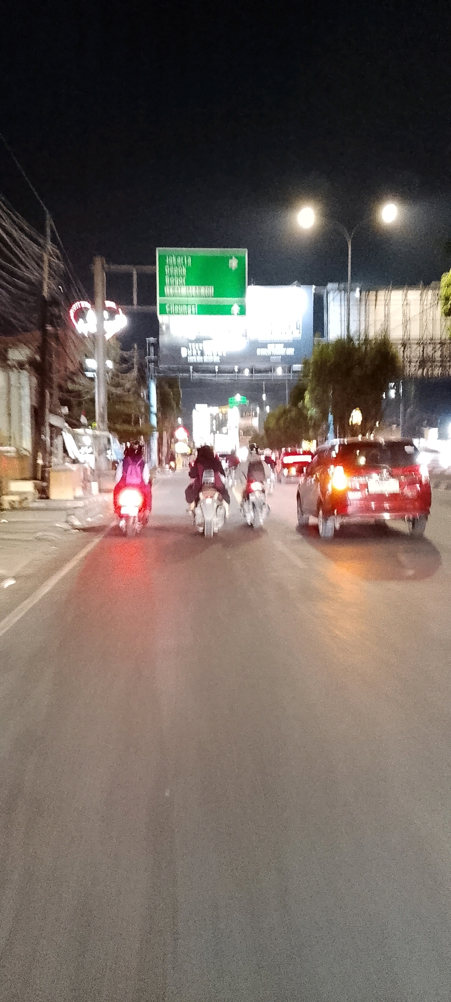 The image depicts a street scene at night in Bekasi, Indonesia, likely on Jalan Ahmad Yani, a major thoroughfare. Multiple motorcycles are visible, with riders wearing helmets and some carrying backpacks. One motorcycle in the foreground has its brake lights illuminated red. A red SUV is also present on the road. Overhead, a large green directional sign indicates "Jakarta" and other destinations. Streetlights illuminate the scene, casting a glow on the asphalt road and the surrounding buildings, which appear to be commercial establishments. Power lines and poles are also visible along the roadside. The overall atmosphere suggests a busy urban environment with active traffic.