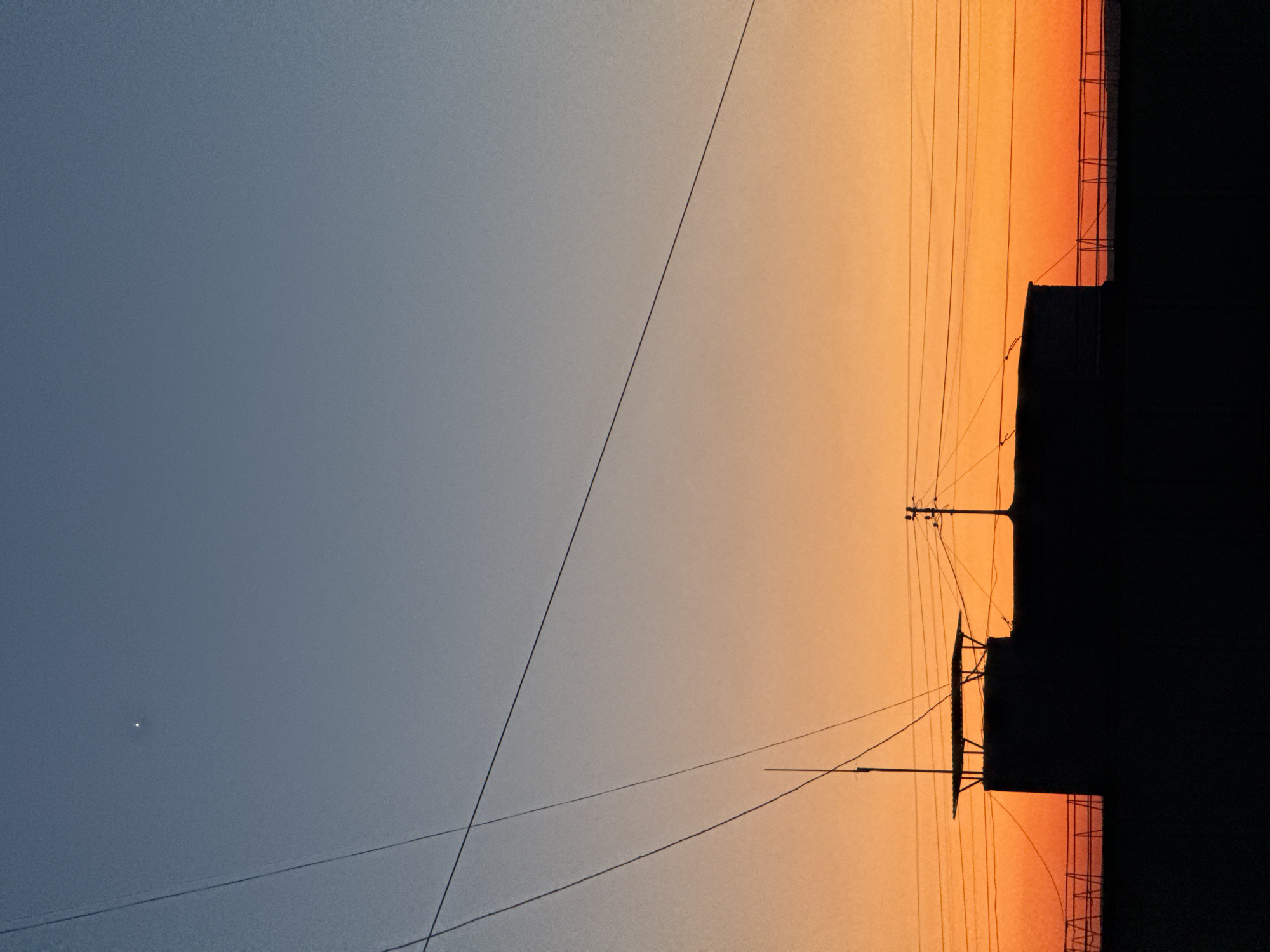 The image shows a view of the sky at dusk in Chaikovsky, Russia. The sky is split between a blueish-grey on the left and a warm orange on the right. Power lines run across the frame. The silhouette of a building and power poles are visible against the orange part of the sky. There is a bright point of light, possibly a star or planet, in the blueish-grey portion of the sky. The image is oriented sideways, with the sky appearing on the left and the ground or built environment on the right.