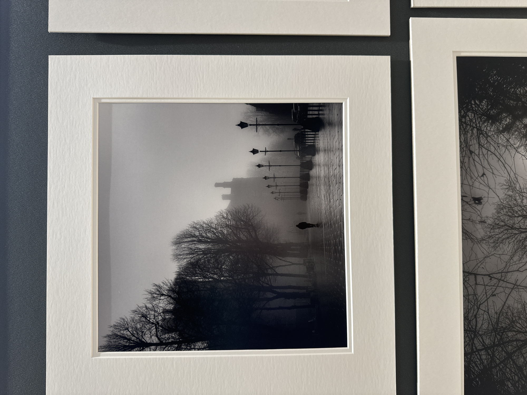The black and white photograph depicts a foggy scene on a cobbled street in Kilkenny, Ireland. The image is framed by a white mat and appears to be part of a larger exhibition, with another matted photograph visible to the right. In the foreground, silhouetted trees with bare branches line the left side of the street. A lone figure, also silhouetted, walks away from the viewer down the wet, reflective street. Several ornate lampposts stand in a row on the right side of the street, their lights muted by the fog. In the background, a building with distinctive chimneys can be seen dimly through the mist. Bicycles are parked against a wall on the left. The overall atmosphere is one of quiet solitude and moody ambiance.