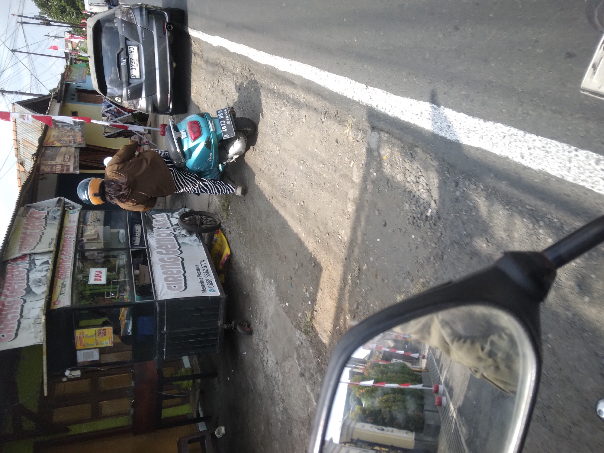 The image captures a street scene in Sokaraja, Indonesia, likely near a food stall. A woman wearing a brown jacket and black and white striped pants is seated on a turquoise scooter. She has a helmet on her head. The scooter has a license plate that reads "B 4672 BBA". To her left is a food cart with the words "CIRENG GEMOY ABA" and a phone number visible. The cart appears to be selling food. A grey Honda Fit car with license plate "R 1737 HJ" is parked on the right side of the road. The road has a white line marking the lane. The environment appears to be a typical street with buildings in the background. The overall impression is a snapshot of daily life on a street in Sokaraja.