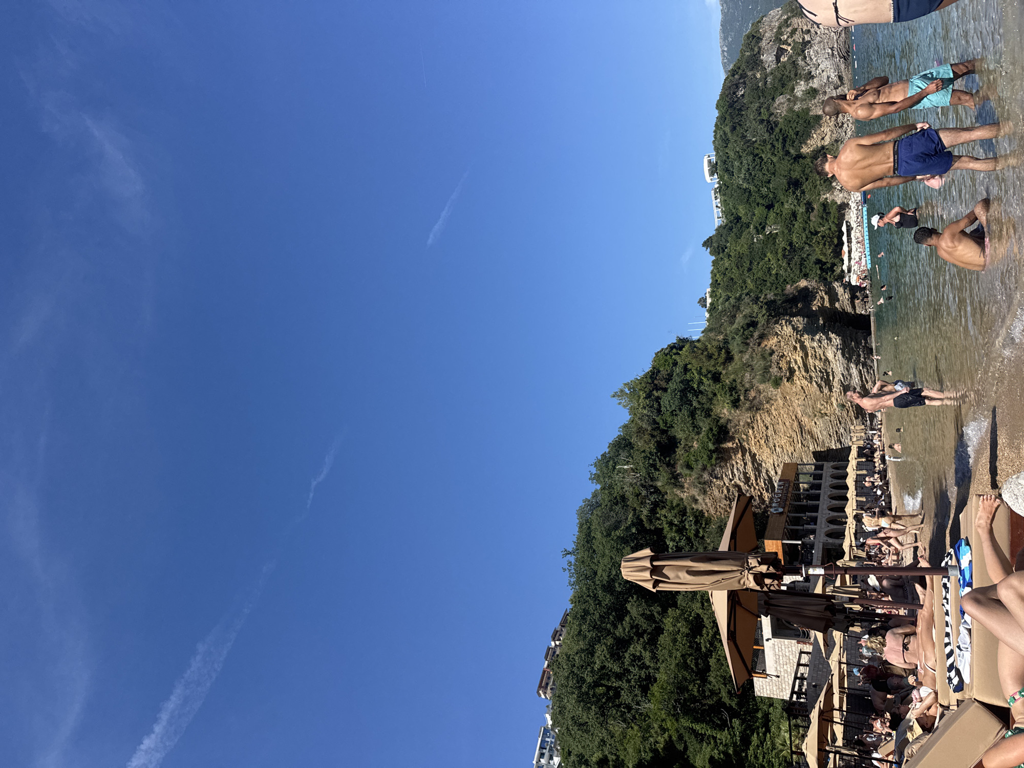 The image captures a sunny day at Mogren Beach in Budva, Montenegro. The foreground shows several people relaxing on beach chairs, some partially visible. Further back, the shallow, clear water of the Adriatic Sea is populated by swimmers. A restaurant with a prominent "MOGREN" sign is visible on the left, with umbrellas and more beachgoers extending down the coastline. The background features a lush, green, tree-covered hillside with buildings perched on top, and a rocky cliff face characteristic of the Budva coastline. The sky is a vibrant blue with a few wispy clouds.