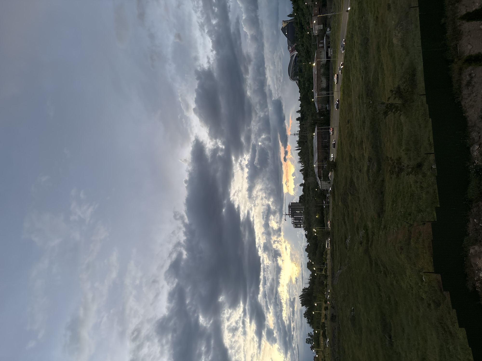 The image depicts a cityscape during a sunset under a cloudy sky. The scene likely captures Yerevan, Armenia, identifiable by the presence of urban structures and the cityscape layout.
The sky is filled with layers of dark, ominous clouds intermixed with patches of lighter, sunset-lit sky displaying orange and yellow hues. The ground consists of a grassy area bordering a road with streetlights, and occasional cars are visible on the road. There are several buildings and structures in the background, including what appears to be a construction site. Along the right edge is a dark boundary which may be a fence or a wall. There are no visible people, and the focus is on the natural sky and urban landscape.