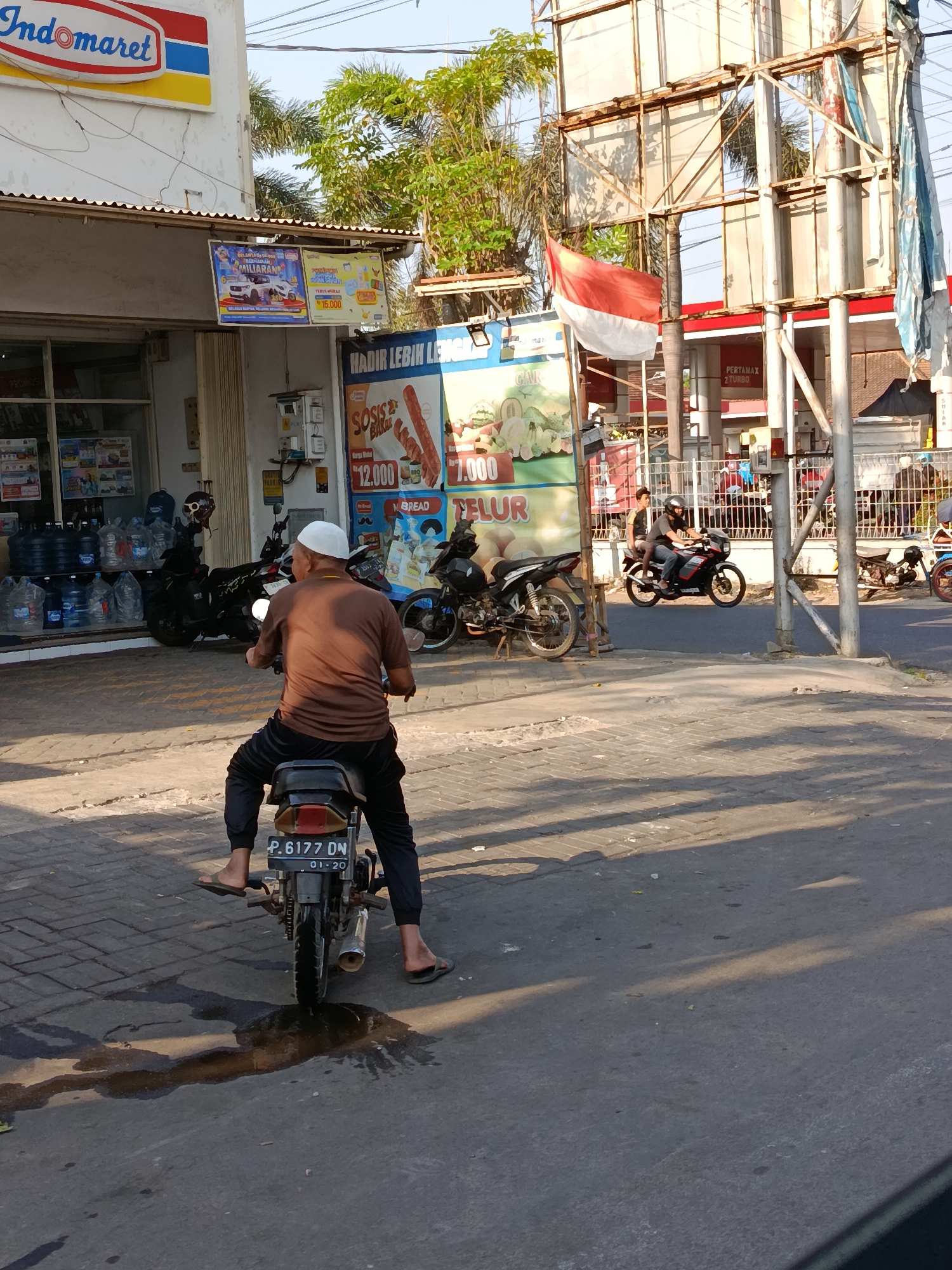 A man wearing a brown shirt and black pants, along with a white skullcap, rides a motorcycle with license plate number P.6177 DN. He is facing away from the camera, looking towards a street scene in Kademangan Kulon, Indonesia. In the background, a red and white Indonesian flag hangs from a metal structure. To the left of the flag, a sign for Indomaret, a convenience store chain, is visible. Adjacent to the Indomaret store, there are several large blue water bottles stacked outside. Other motorcycles are parked and in motion on the street. The ground is paved with interlocking concrete blocks in some areas and asphalt in others, with some wet patches visible on the asphalt. The overall lighting suggests it is daytime, possibly late afternoon due to the shadows.
