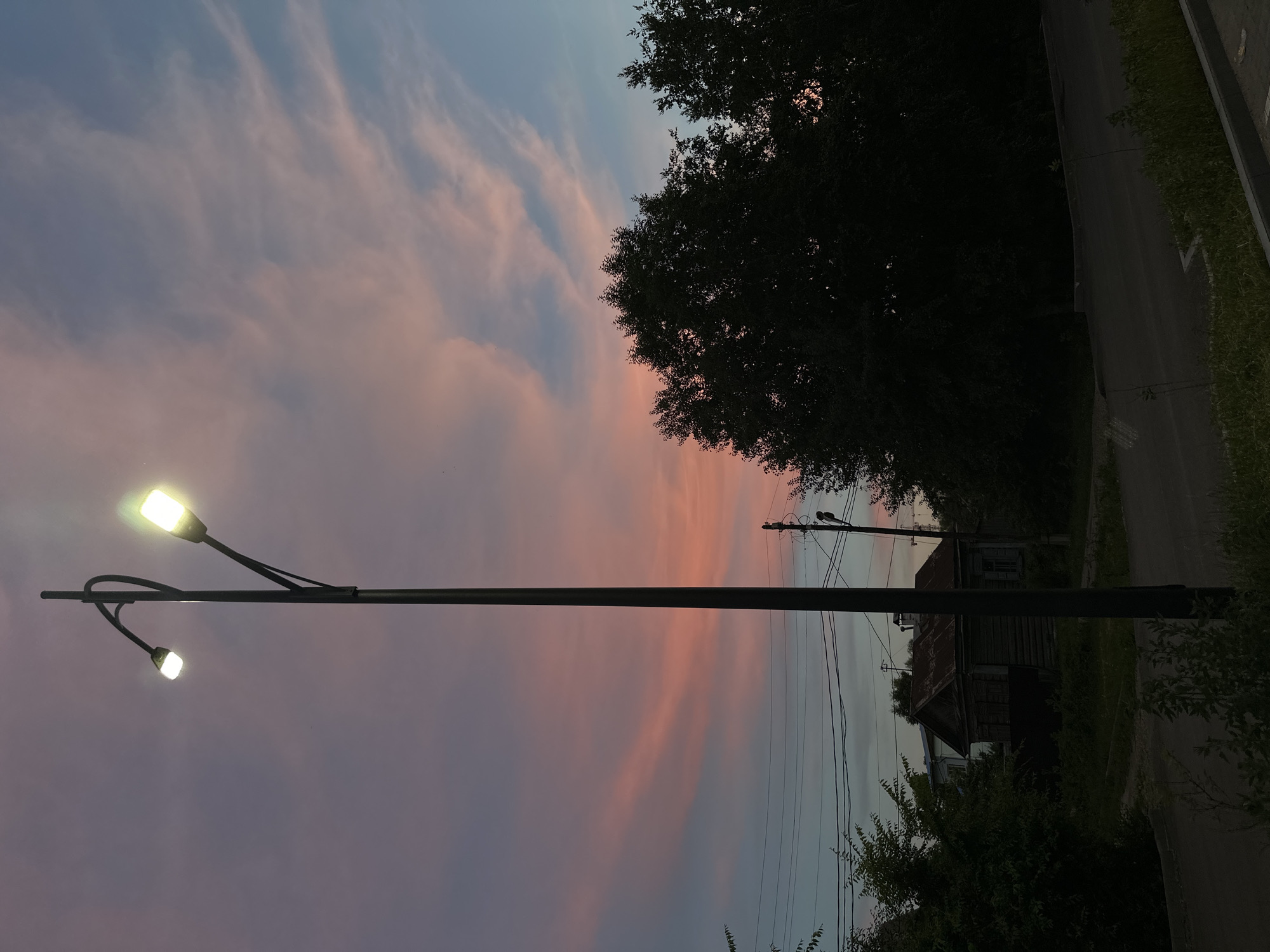 The image captures a dusk scene in Blagoveshchensk, Russia, with a prominent black lamppost with two illuminated lamps standing in the foreground. The sky displays a gradient of colors, transitioning from a soft pink and purple hue, indicative of sunset, to a muted blue towards the horizon. Wispy clouds are scattered across the sky, catching the light and enhancing the atmospheric effect.
In the middle ground, a small, rustic wooden house with a weathered roof is visible, partially obscured by dark green foliage. Power lines stretch across the background, supported by poles, suggesting an urban or semi-urban setting. Dense trees with dark silhouettes are positioned on the right side of the frame, further emphasizing the fading daylight. A paved road and grassy areas are present in the lower portion of the image, with a concrete curb defining the edge of the road. No people or animals are depicted in the scene, and no discernible actions are taking place. The overall atmosphere is tranquil and serene, characteristic of twilight.
