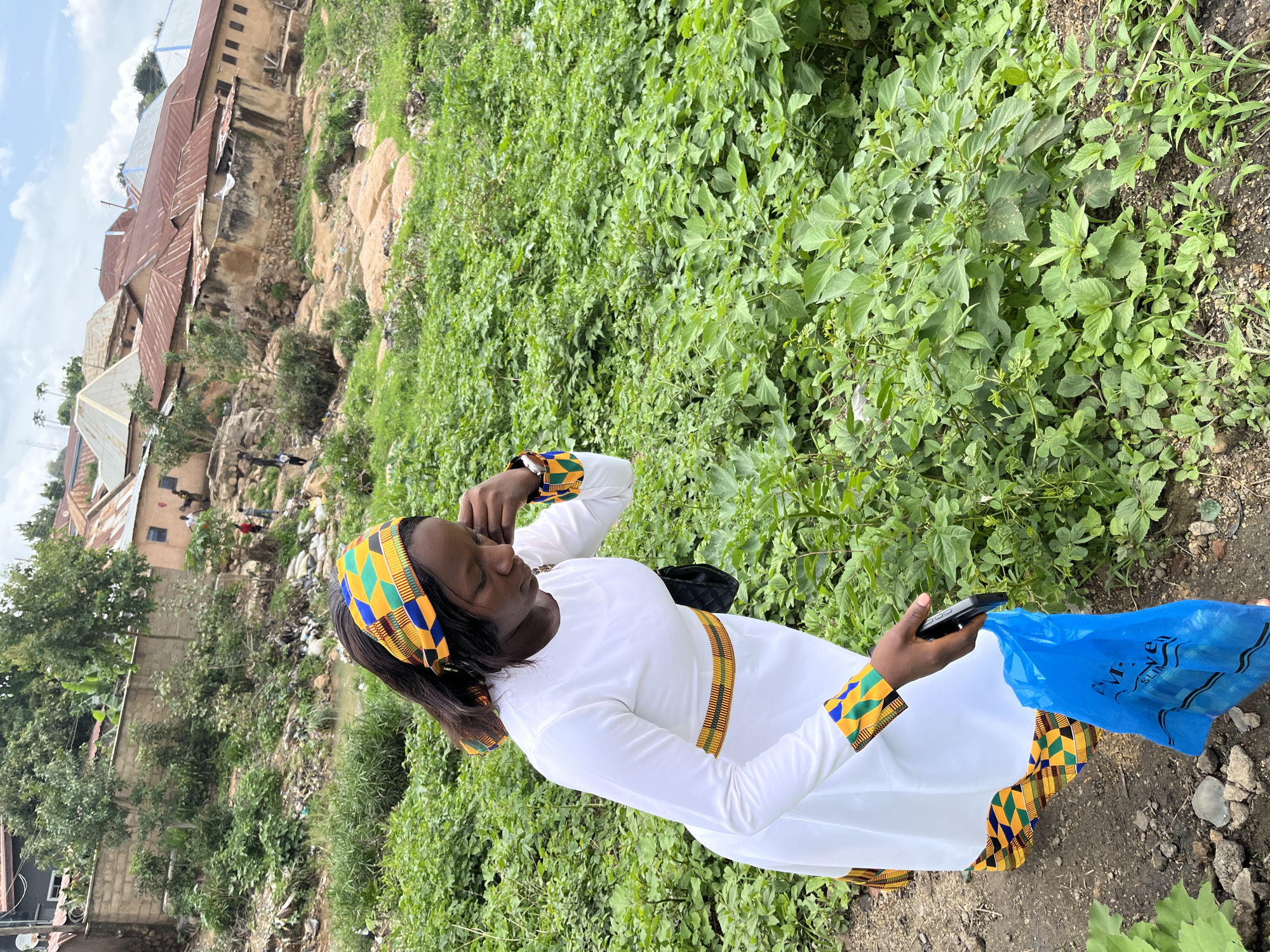 A woman stands on a dirt path, angled slightly towards the right of the frame, within a semi-urban or informal settlement in Jos, Nigeria. She wears a white long-sleeved dress featuring a patterned band of colorful geometric fabric (yellow, blue, green, black, red) at the waist, cuffs, and hemline. A matching headband adorns her dark hair. Her gaze is directed downwards and to her right, with her right hand raised near her ear. In her left hand, she holds a black smartphone and a blue plastic bag with white text. A black shoulder bag strap is visible over her left shoulder.
The immediate environment to her right and extending into the mid-ground consists of dense, lush green vegetation covering an uneven terrain. The background reveals a steep hillside dotted with numerous residential structures. These buildings exhibit varied construction, primarily featuring earth-toned walls and a predominance of rusted, corrugated metal roofs, interspersed with some newer, silver-colored roofs. A narrow, dark water channel or drainage ditch, containing visible litter, runs along the base of the hillside. Further up the slope, several small figures, likely people, are discernible navigating the paths between the houses. Trees and sporadic vegetation are present throughout the background. The sky above is partly cloudy.