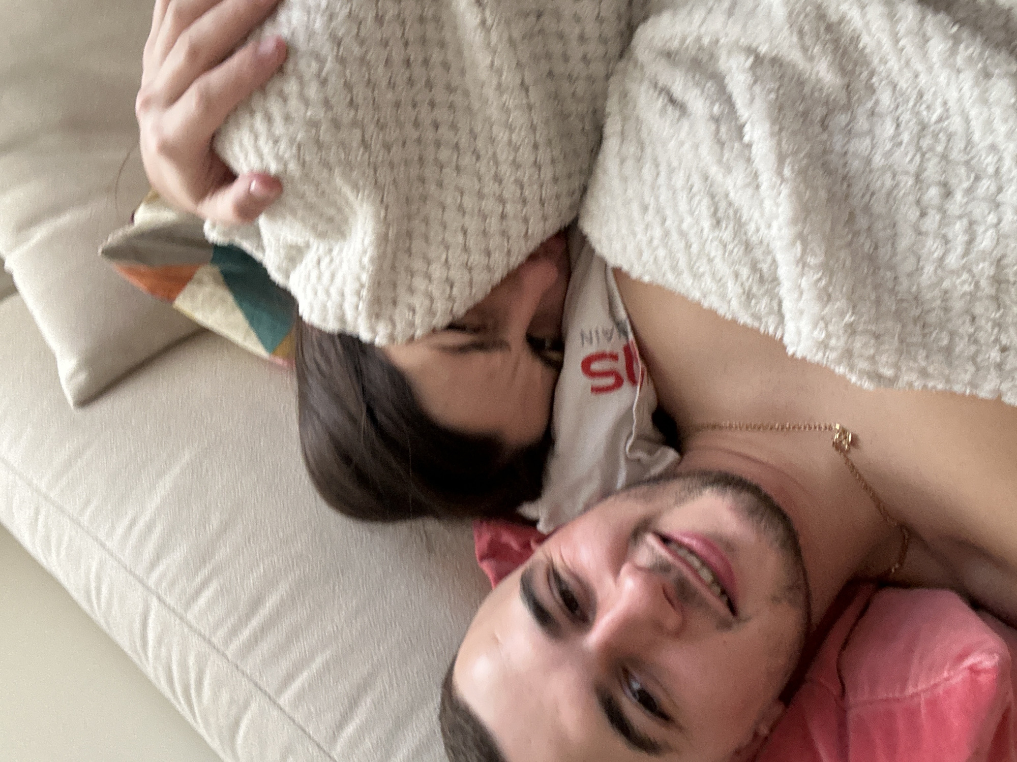 A man and woman are lying on a light beige sofa. The woman has dark hair and is partially covered by a white blanket. The man is smiling at the camera, wearing a pink shirt, and has a gold necklace. A pillow with geometric patterns is visible next to the woman's head. The location of the photo is unavailable; there is no evidence it was taken in Plovdiv, Bulgaria.