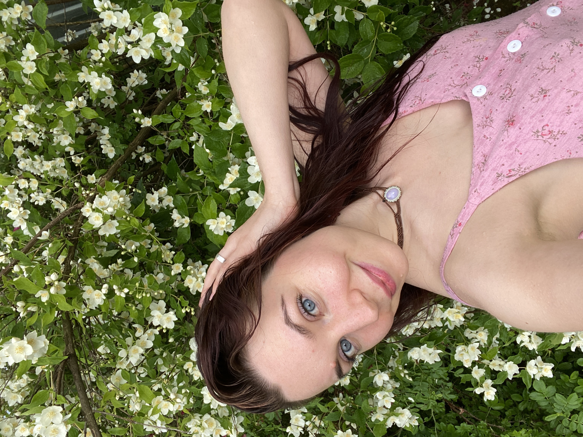 A young woman with dark brown hair and blue eyes lies in a bed of white jasmine flowers. She is wearing a pink floral-print dress with white buttons and a brown string necklace with a light purple stone pendant. Her right hand is behind her head, and her left arm is resting on the flowers. She is looking up at the camera with a soft smile. The scene appears to be outdoors, likely in a garden or park, with abundant greenery and white blossoms. The lighting suggests it is daytime, possibly late afternoon or early evening due to the soft light. The overall mood is serene and peaceful. No visible text is present in the image.