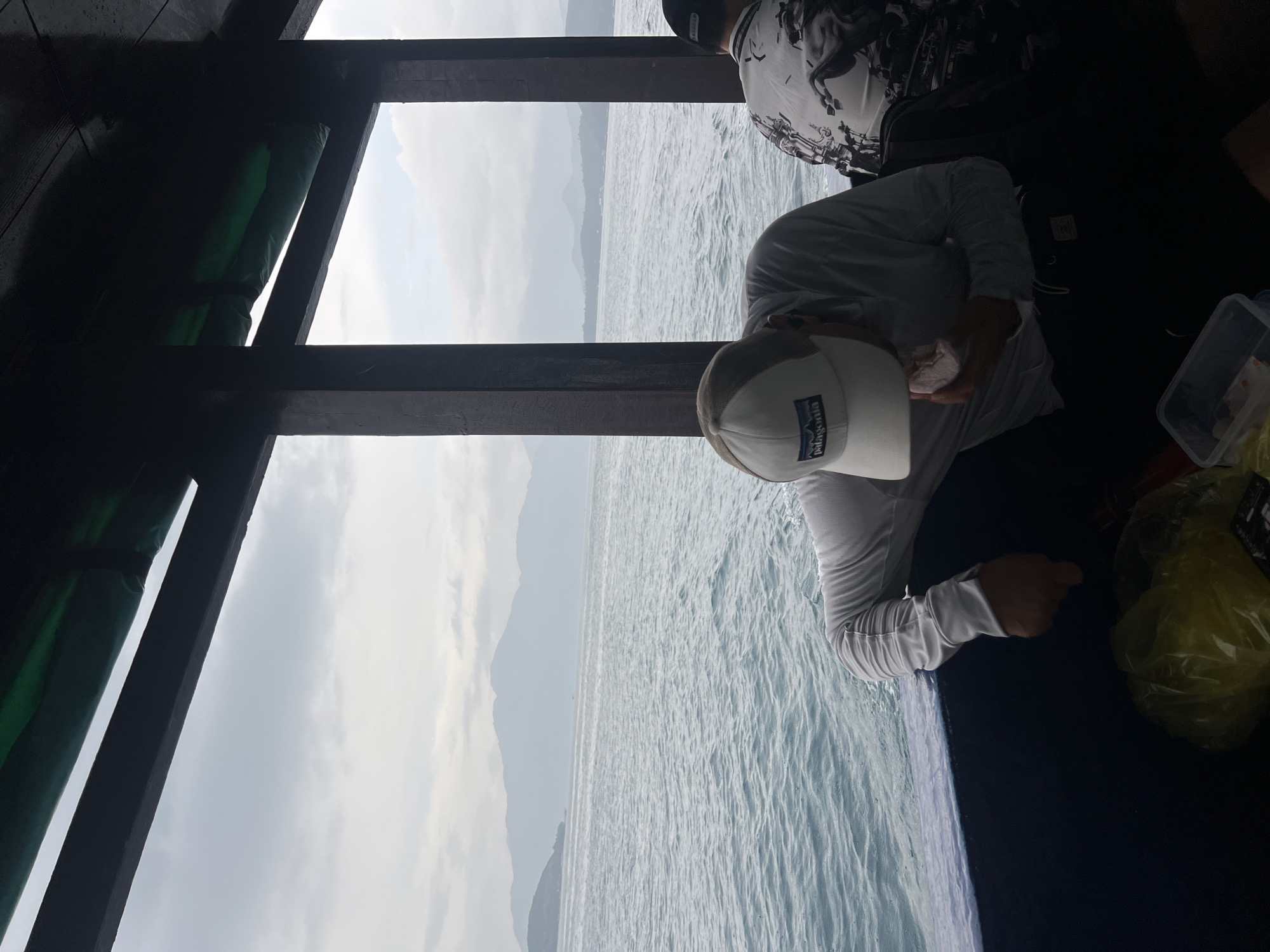 The image shows a view from inside a boat, possibly in the sea near Bac Nha Trang Ward, Vietnam. The sea and distant mountains are visible through the boat's frame. A person wearing a white shirt and a baseball cap with a "Patagonia" logo is seated, appearing to eat something from a piece of paper. Another person, partially visible in a patterned shirt, is sitting nearby. A clear plastic container and a yellow plastic bag are also present near the seated individual.