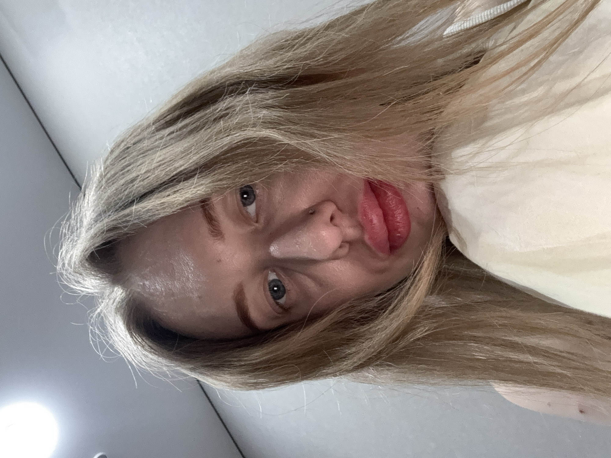 An indoor self-portrait depicts a female individual, asserted to be located in Rostov-on-Don, Russia. The subject has long, light blonde hair, which appears loose and partially obscures the face and shoulders. Her face is viewed from a low angle, with blue-grey eyes looking directly at the camera. A small silver stud piercing is visible in the left nostril. Her lips are full, enhanced with red/pink lipstick, and appear slightly parted or puckered. She wears a light-colored, possibly cream or light yellow, garment over her right shoulder and chest, with the left shoulder appearing bare. The background consists of plain, light-colored walls, with a bright circular overhead light source present in the upper left quadrant of the image.