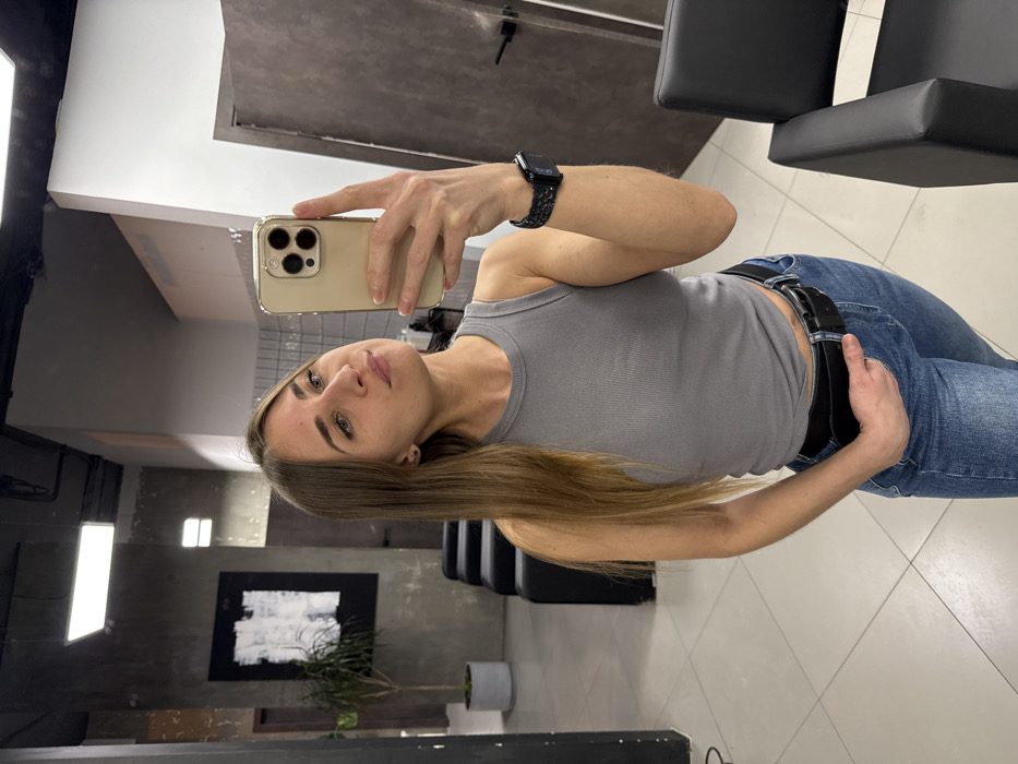 A woman with long blonde hair poses in a mirror selfie, holding a gold-colored smartphone. She wears a grey tank top, blue jeans with a black belt, and an Apple Watch on her left wrist. The background shows a salon interior with salon chairs, a painting, and a potted plant. The floor has a tile pattern. The lighting appears bright, highlighting the woman's features.