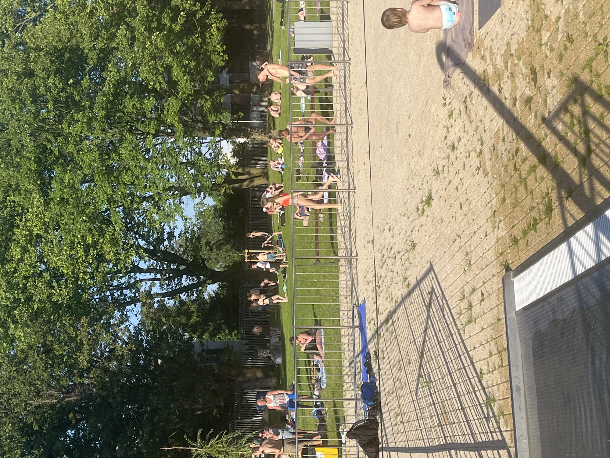 The image depicts a sunny, outdoor scene, likely at a public park or recreational area. The time of day appears to be late afternoon, given the long shadows cast by the fence and trees. The weather is warm and clear, suitable for outdoor activities.
The primary subjects are a group of people engaged in various leisure activities. Many are sunbathing on a grassy area, while others are seen interacting or relaxing. The people are dressed in casual summer attire, including swimsuits and shorts. One notable individual is a young child in a diaper, lying on a towel.
The scene is divided by a metal fence, which runs horizontally across the image. On one side of the fence, there is a well-maintained lawn with several trees providing shade. On the other side, there is a sandy or gravel area with some sparse vegetation. A part of a building with white siding and a dark grey reflective surface is visible at the far right edge of the frame, suggesting it might be part of the facility.
The overall mood is relaxed and leisurely. People appear to be enjoying the warm weather and the amenities of the location. There is no visible text in the image.