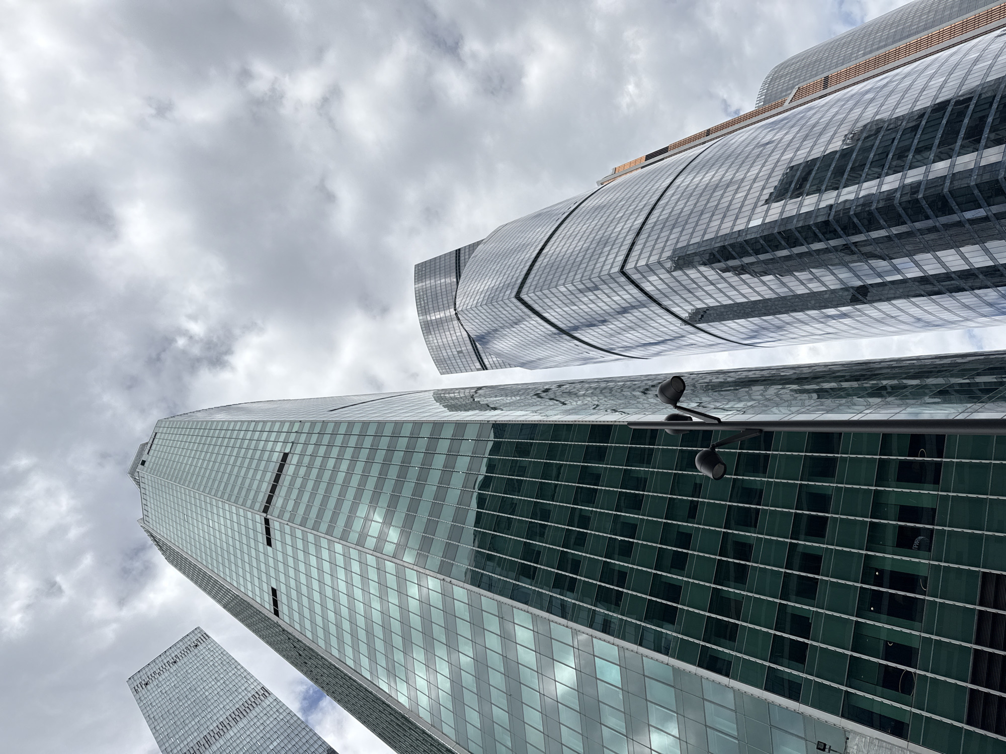 The image depicts a low-angle view of several skyscrapers against a cloudy sky. The buildings are modern in design, with glass facades. One building has a distinctive curved shape. A street lamp is visible in the foreground. The location is likely a dense urban area in a city, but the specific location is not identifiable.