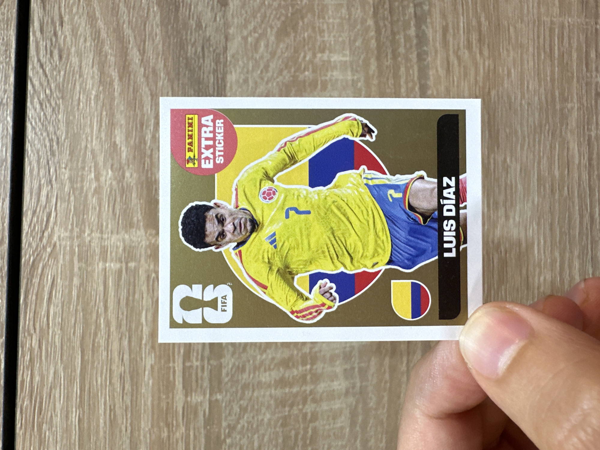The image shows a soccer sticker featuring Luis Díaz. The sticker has a gold background and displays the FIFA logo on the left and the Panini "EXTRA STICKER" logo on the upper right. Luis Díaz is depicted in a yellow jersey with red and blue accents, and blue shorts. He is wearing the number 7. The Colombian flag is visible on the sticker. The sticker is held by a hand in the lower left corner of the image, with the person's thumb and forefinger obscuring a small portion of the sticker. The background of the image is a wooden surface. There is no information in the image that relates to the city of Prisovo, Bulgaria.