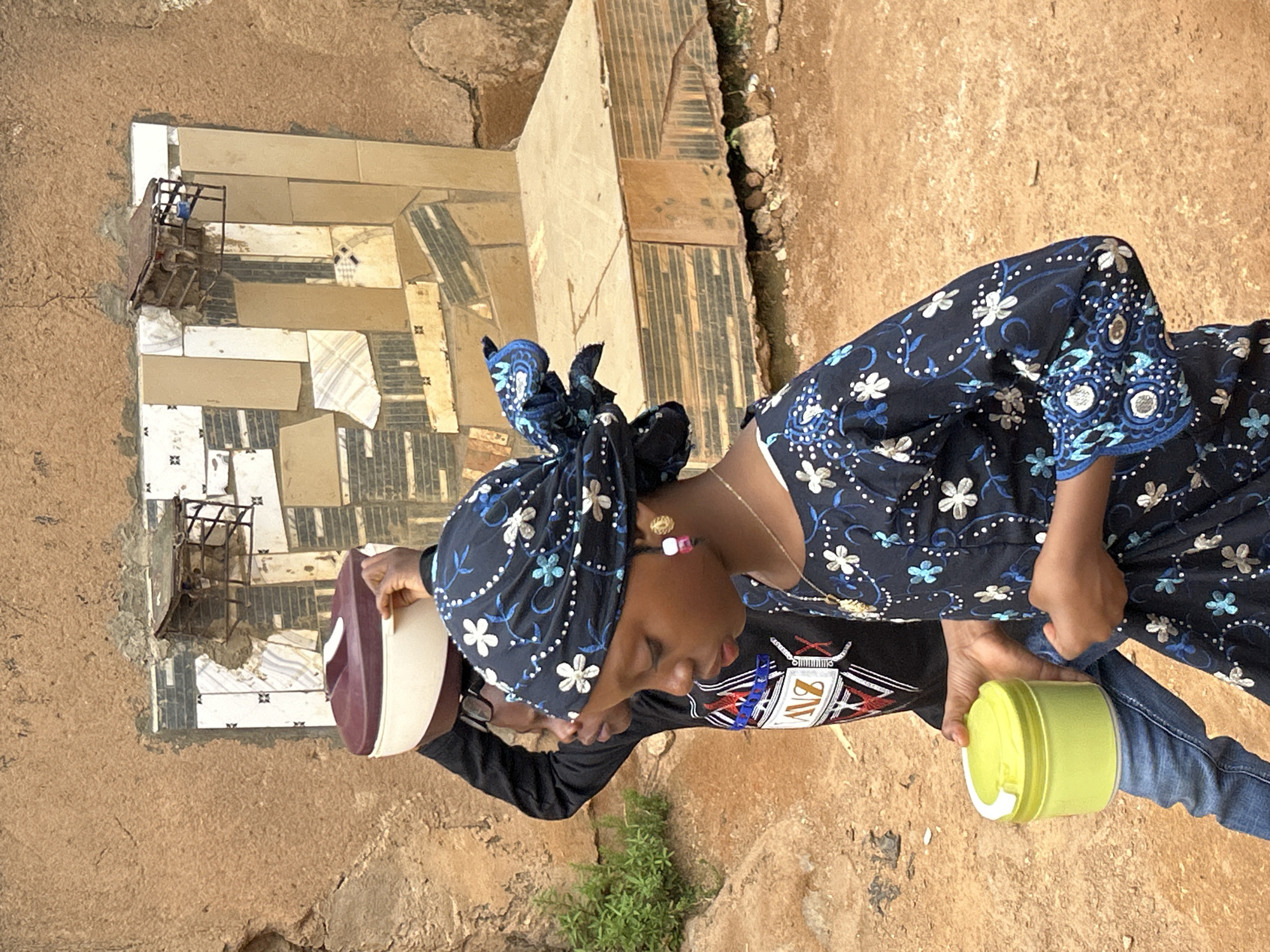 A person with dark skin wearing a blue floral headscarf and matching dress stands outside, likely in Jos, Nigeria, holding a maroon and white container in their left hand and a lime green container in their right. The person's head is bowed, and they wear gold earrings and a necklace. A second person, wearing a black long-sleeved shirt with a colorful graphic, is visible behind them, partially obscured, holding the maroon and white container. The ground is dirt, with some sparse green vegetation visible in the foreground. Behind the figures, there is a partially tiled structure, possibly a low wall or step, featuring a mix of beige, brown, and white tiles. A metal frame structure rests against the tiled surface. The background consists of a dirt wall.