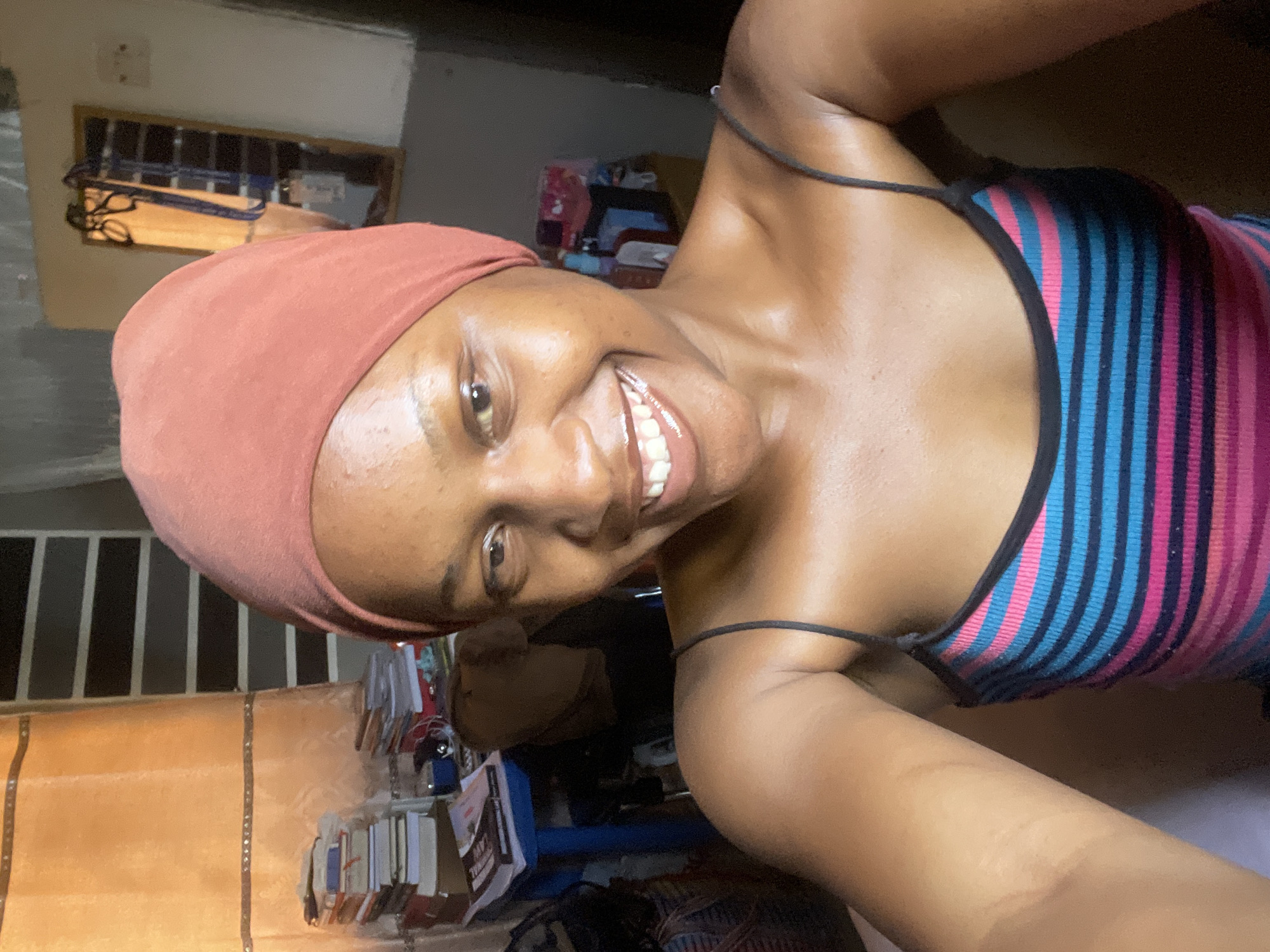 A woman is taking a selfie in a room, with a pink headscarf and a striped top. She is smiling broadly and her eyes are open, suggesting a happy or amused emotion. The background of the image is cluttered with various objects, including books, what appear to be clothes, and possibly some shelves or furniture. The lighting suggests it might be indoors, possibly at night or in a dimly lit room. There is no clear indication of the weather or specific time of day, other than the indoor setting. The location context is Jos, Nigeria, but there are no specific visual cues in the image to confirm this. There is no visible text in the image.
