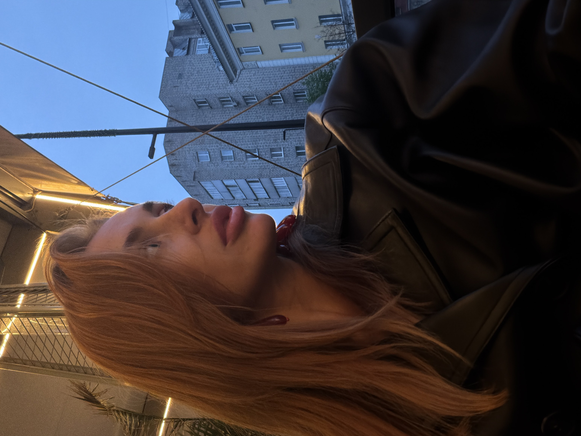 The image shows a woman with long, auburn hair wearing a black leather jacket. Her face is in the foreground, with her lips and nose clearly visible. In the background, there is a multi-story building with many windows, suggesting an urban environment. The presence of overhead wires and a partially visible structure with lighting indicates that the photo may have been taken in an outdoor cafe or restaurant setting. Based on the architecture of the building and the overall urban landscape, it is likely that the location is within the city of Krasnoyarsk, Russia.