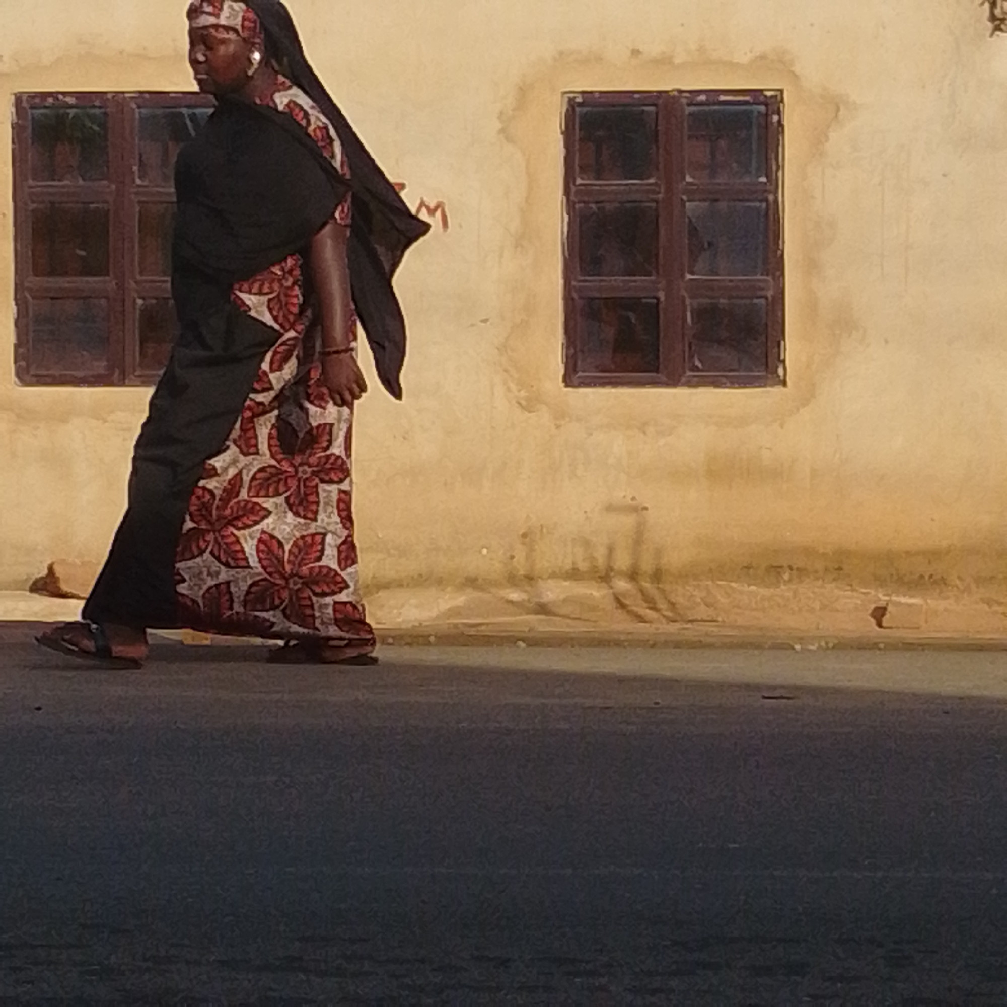 A person walks in front of a yellow building with two dark-paned windows. The person is wearing a black headscarf and a black and red floral patterned dress. They are also wearing sandals. The scene appears to be outdoors, possibly on a street. The building has a rough texture, and there are some faint markings or graffiti on its surface. The lighting suggests it is daytime, with shadows cast on the ground. The overall impression is of a simple, everyday scene in a location that appears to be in Nigeria, possibly in Tempure, as indicated by the location context. No specific activity or event is clearly depicted beyond the act of walking. The image is somewhat grainy, which could be due to low light conditions or the resolution of the original photograph. The person's expression is not clearly visible.