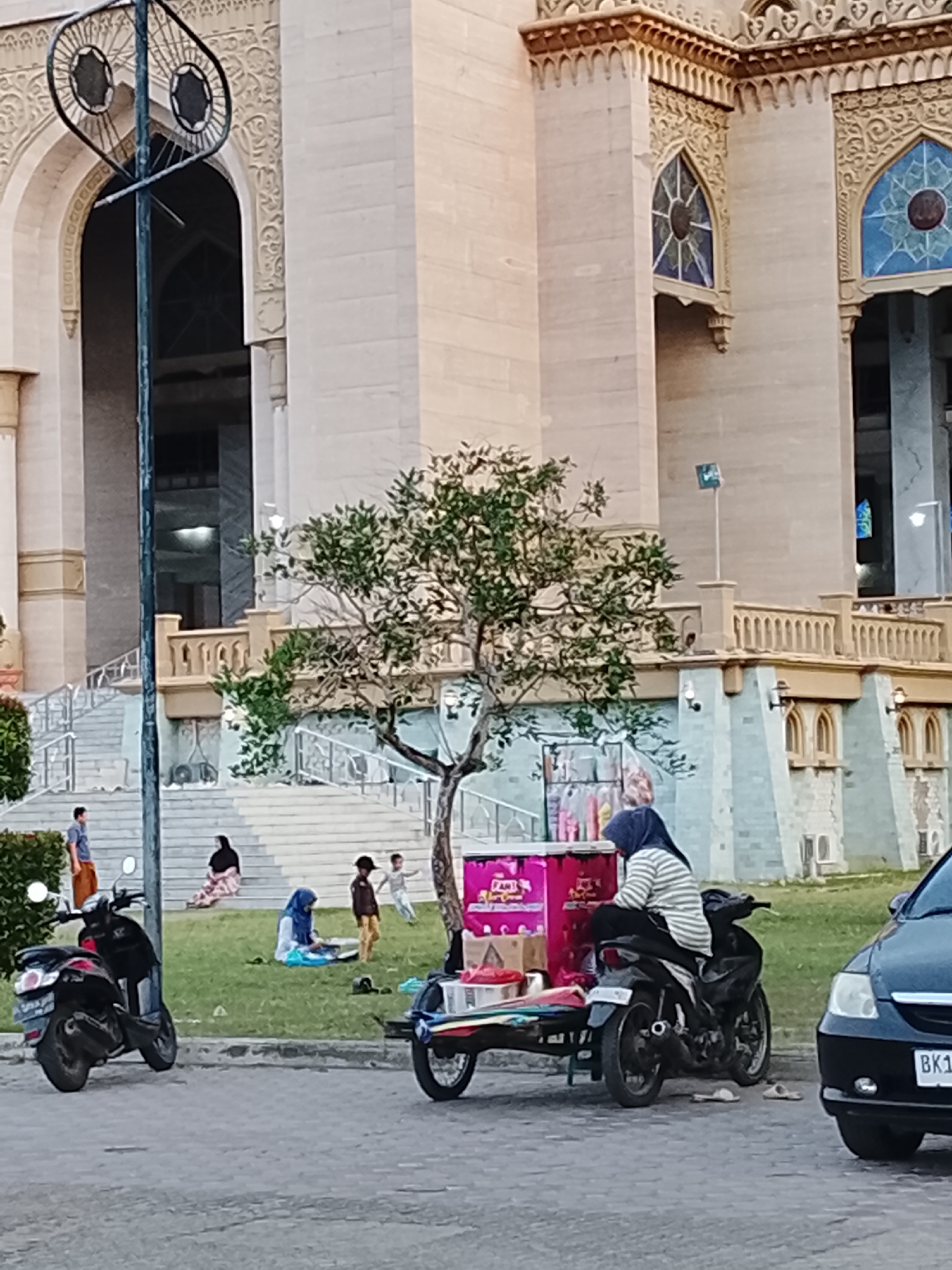 The image depicts the exterior of a large, ornate building, likely a mosque, with intricate architectural details, including arched doorways and decorative window frames. In front of the building, a group of people are gathered on a grassy area. To the left, a black motorcycle is parked. In the center, a woman wearing a headscarf is seated on a motorcycle attached to a small cart, which appears to be a food or beverage stand. The cart is brightly colored with pink and white accents. Several people, including children, are visible in the background, some sitting on stairs and others playing on the grass. A black car is parked on the right side of the image. The scene suggests a public space within the city of Lhokseumawe, Indonesia, potentially a gathering place or tourist attraction.