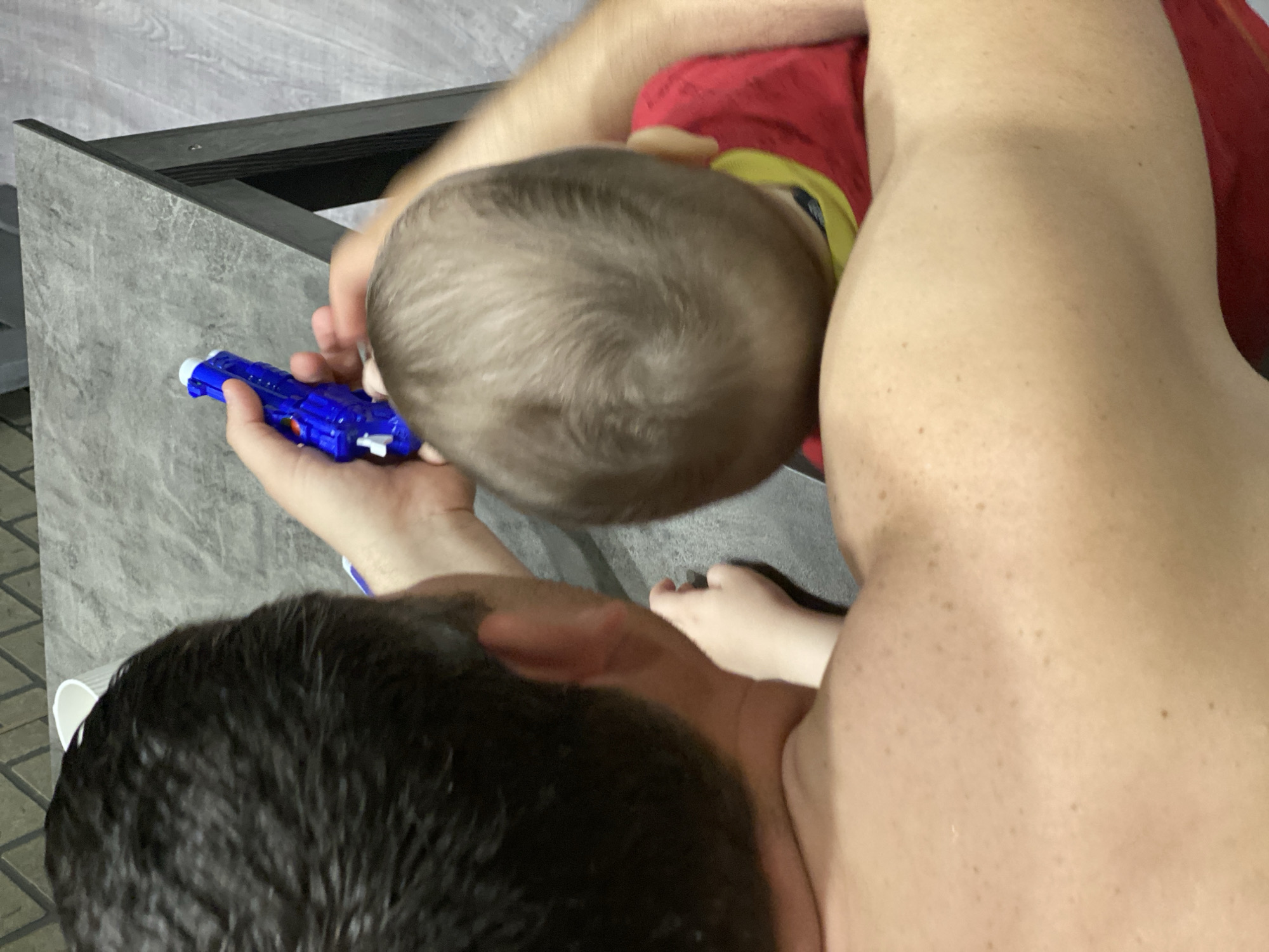 An overhead view captures two individuals in an indoor setting in Sochi, Russia. In the foreground, the bare back and right shoulder of an adult, presumed male, with dark, closely-cropped hair and numerous small freckles/moles, is visible. The adult's right hand holds a bright blue plastic toy gun with white accents. Positioned in front of the adult, partially obscured, is a young child, likely an infant or toddler, with light brown hair and wearing a red shirt with yellow trim. A small hand, presumably belonging to the child, rests near the adult's right ear/neck area. The individuals are positioned adjacent to a dark gray, textured tabletop. A white cup is partially visible on the table. The background features a dark gray wall with a subtle geometric pattern and a light gray floor, possibly wood laminate or tile. The scene depicts an interaction between the adult and child involving the toy.