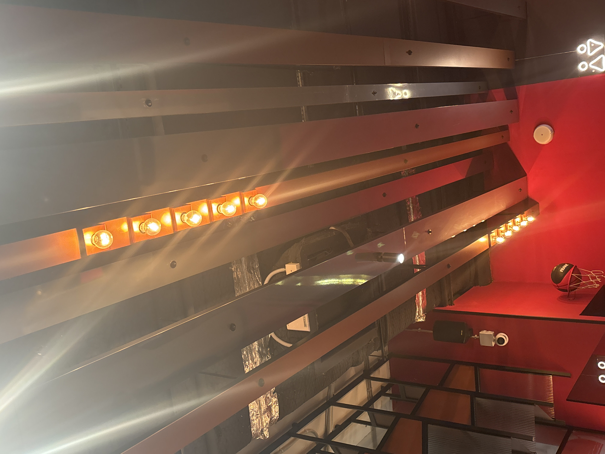 The image shows an indoor space with a red wall on the right side of the frame. A series of parallel, linear light fixtures, some with illuminated bulbs, are arranged horizontally across the ceiling. Below, there is a shelf or counter with a basketball and a net on top of it. There is a set of men and women bathroom signs. There is a surveillance camera attached to the wall. Some metallic support structures are shown below. The location is Unknown, Russia.