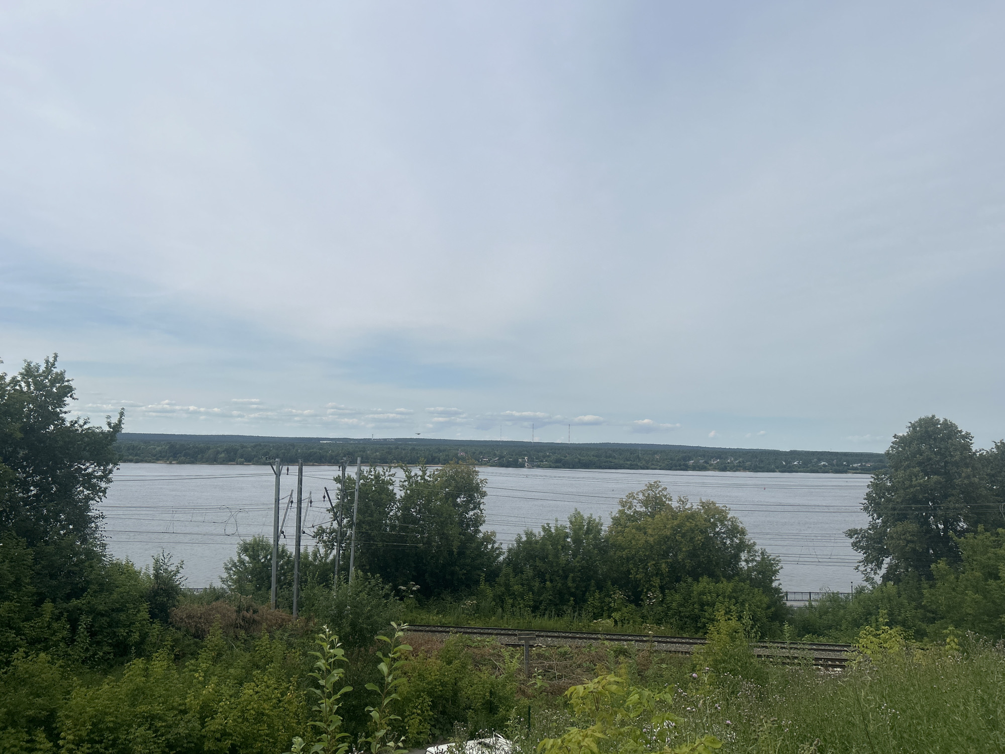 A wide, tranquil river stretches across the horizon under a pale, overcast sky. Along the riverbank, a dense line of green trees forms a natural border, with some taller trees punctuating the landscape. In the foreground, lush green foliage and bushes dominate the scene, with a set of railway tracks running horizontally across the middle ground. Several utility poles with wires are visible between the trees and the river, suggesting the presence of electrical infrastructure. The scene appears to be a natural landscape within or on the outskirts of the city of Perm, Russia, potentially offering a view of the Kama River.
