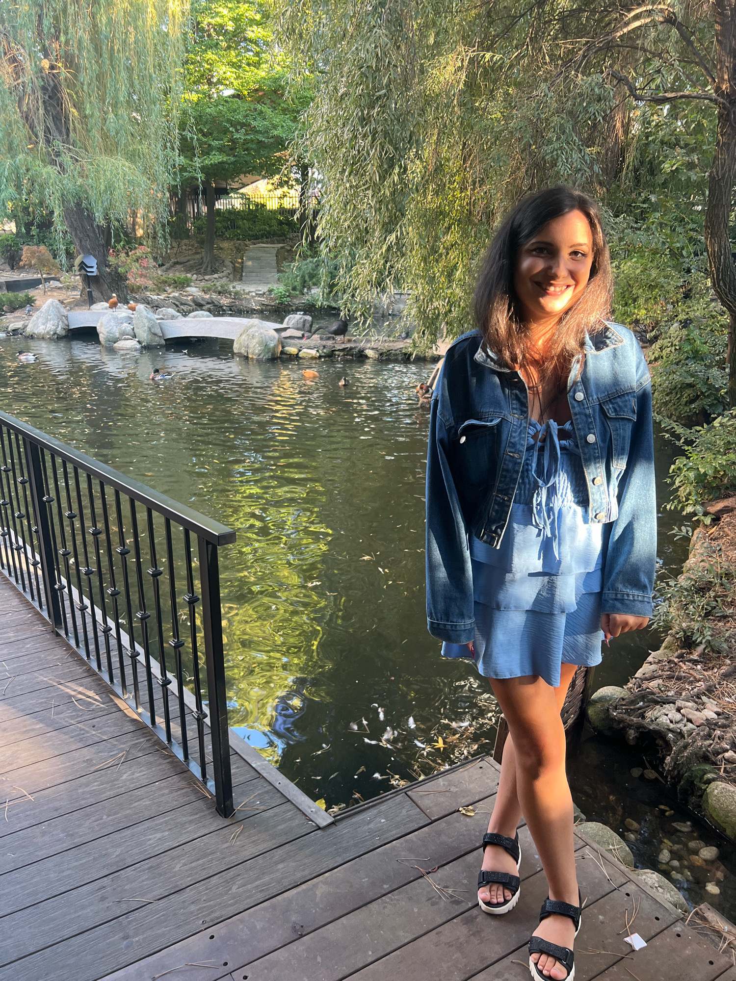 A young woman with dark hair and a bright smile stands on a wooden deck beside a pond. She is wearing a denim jacket over a light blue tiered dress and black sandals with white soles. The pond is surrounded by rocks and lush greenery, with a small arched bridge visible in the background. Several ducks are swimming in the water. The scene appears to be in a park or garden setting. The specific location in Sofia, Bulgaria is not determinable from the image alone.