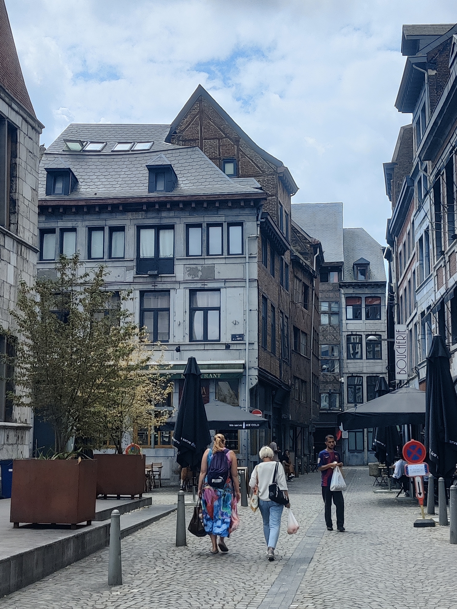 The image depicts a street scene in the city of Liège, Belgium, likely within the historic center, given the architectural style of the buildings. The street is paved with cobblestones. Several people are walking along the street, some carrying shopping bags. One woman in the foreground is wearing a colorful dress and carrying a backpack. Another woman is wearing a white top and jeans. A man in a blue and red striped shirt is also walking, holding plastic bags. The buildings lining the street are multi-story structures with traditional European architecture, featuring mansard roofs, dormer windows, and ornate facades. Some buildings appear to house businesses, with signage visible, including "BOUCHERIE" on one building. Outdoor seating with umbrellas is set up in front of some establishments, suggesting a cafe or restaurant setting. The sky is cloudy. The overall atmosphere is that of a typical day in a European city.